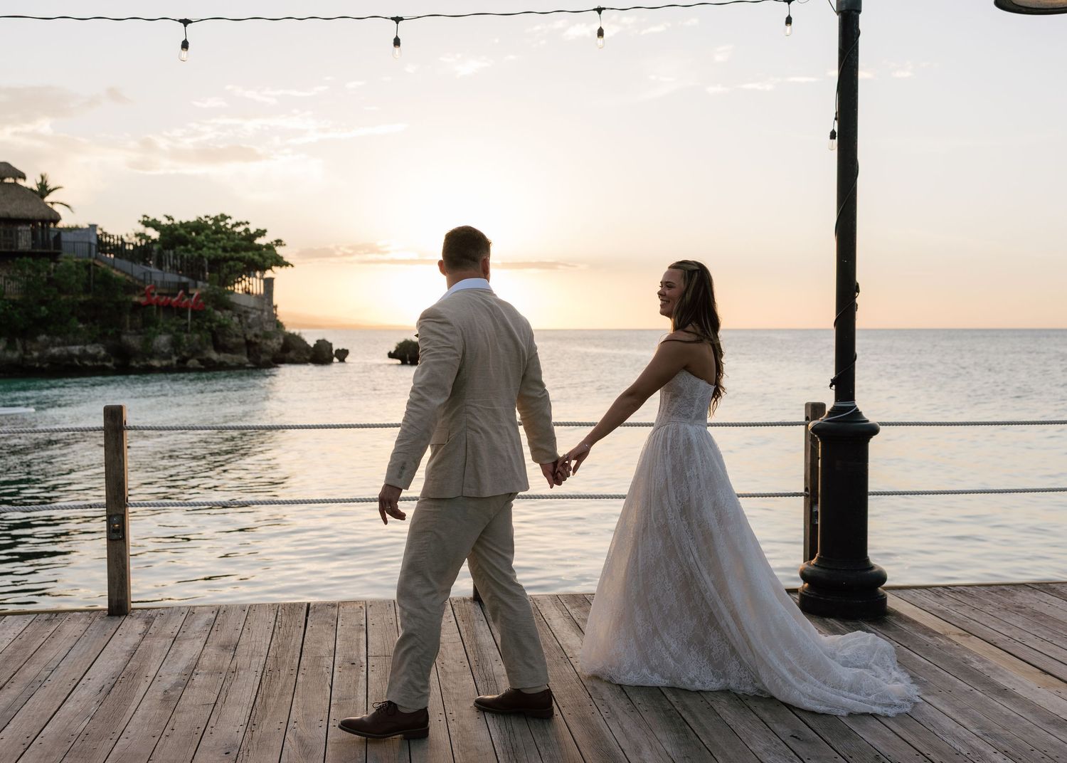 Bride and groom holding hands on a wooden dock at sunset overlooking the ocean at a tropical wedding venue.