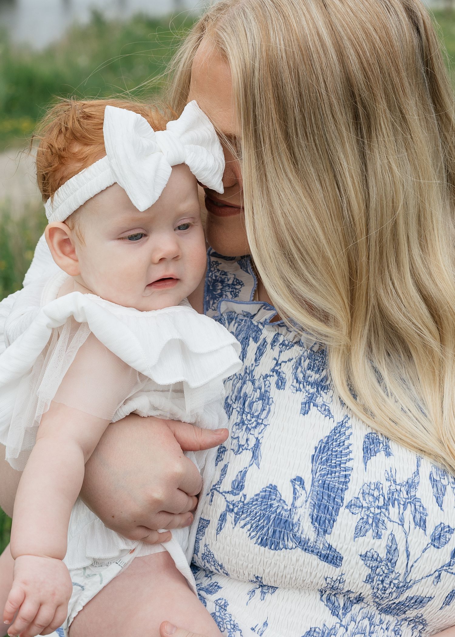 A blonde mother tenderly kisses her red-haired baby girl in a white bow headband and ruffled dress outdoors.