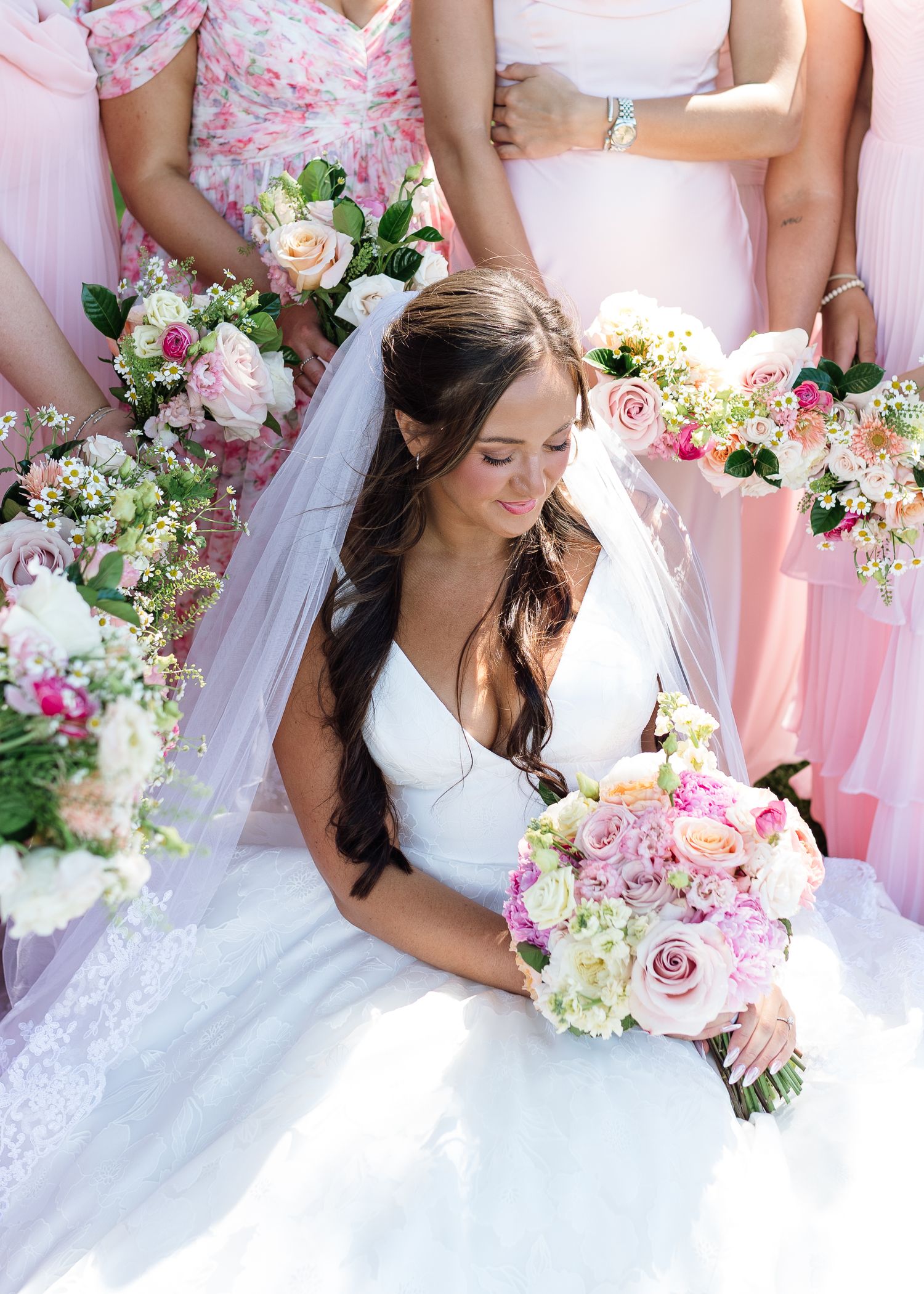 Bride in white gown with veil holds pink rose bouquet surrounded by bridesmaids in pink dresses.