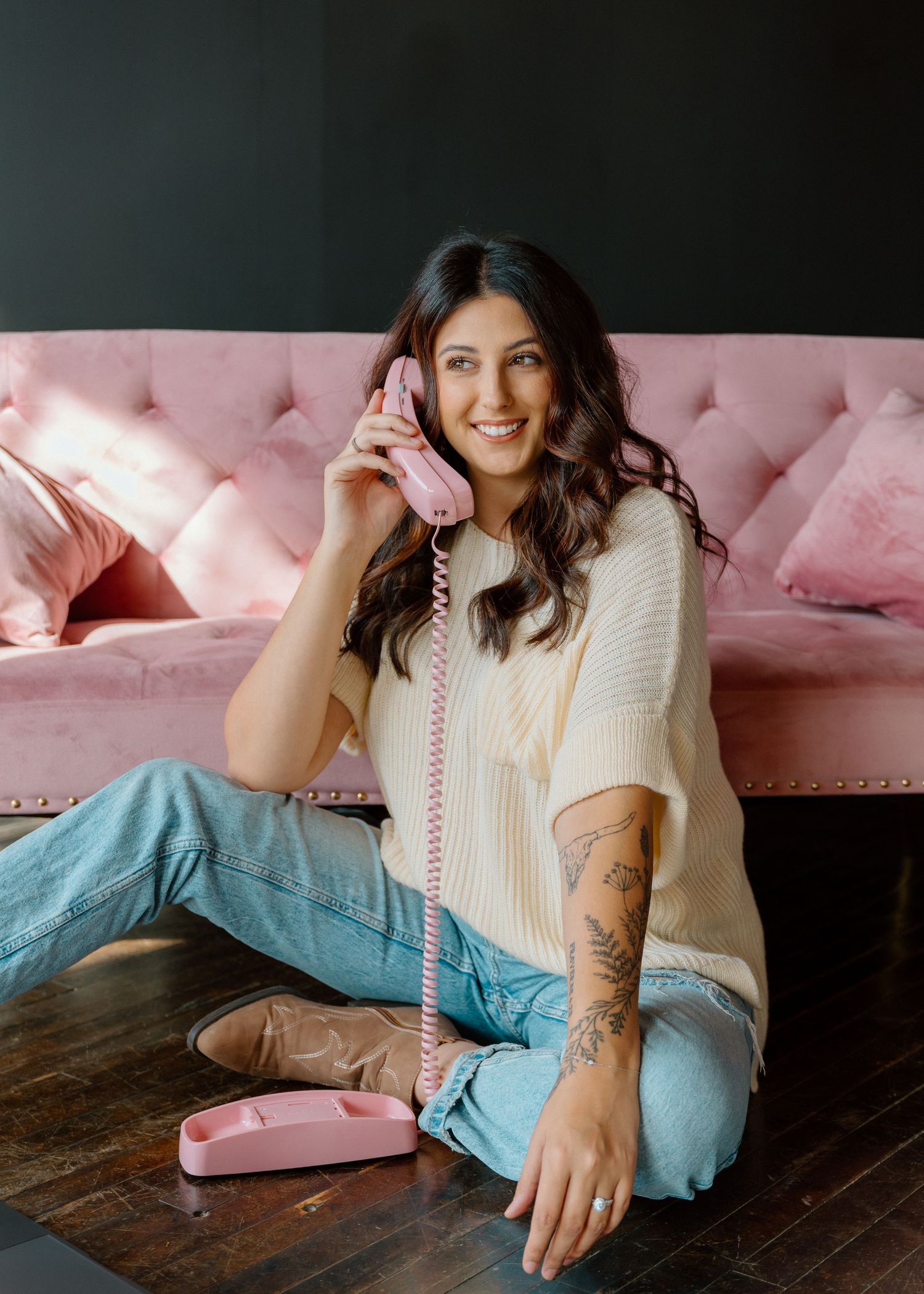 Woman in white sweater and blue jeans lounges on pink cushions against rustic wooden floor.