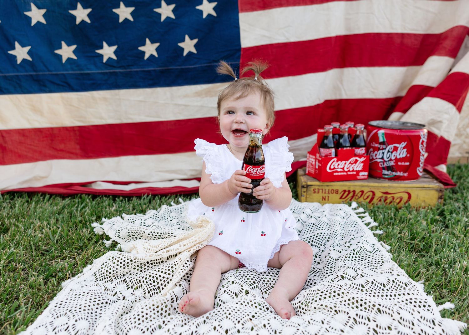 Baby girl in white romper sits on lace blanket holding Coca-Cola bottle before American flag on Fourth of July.