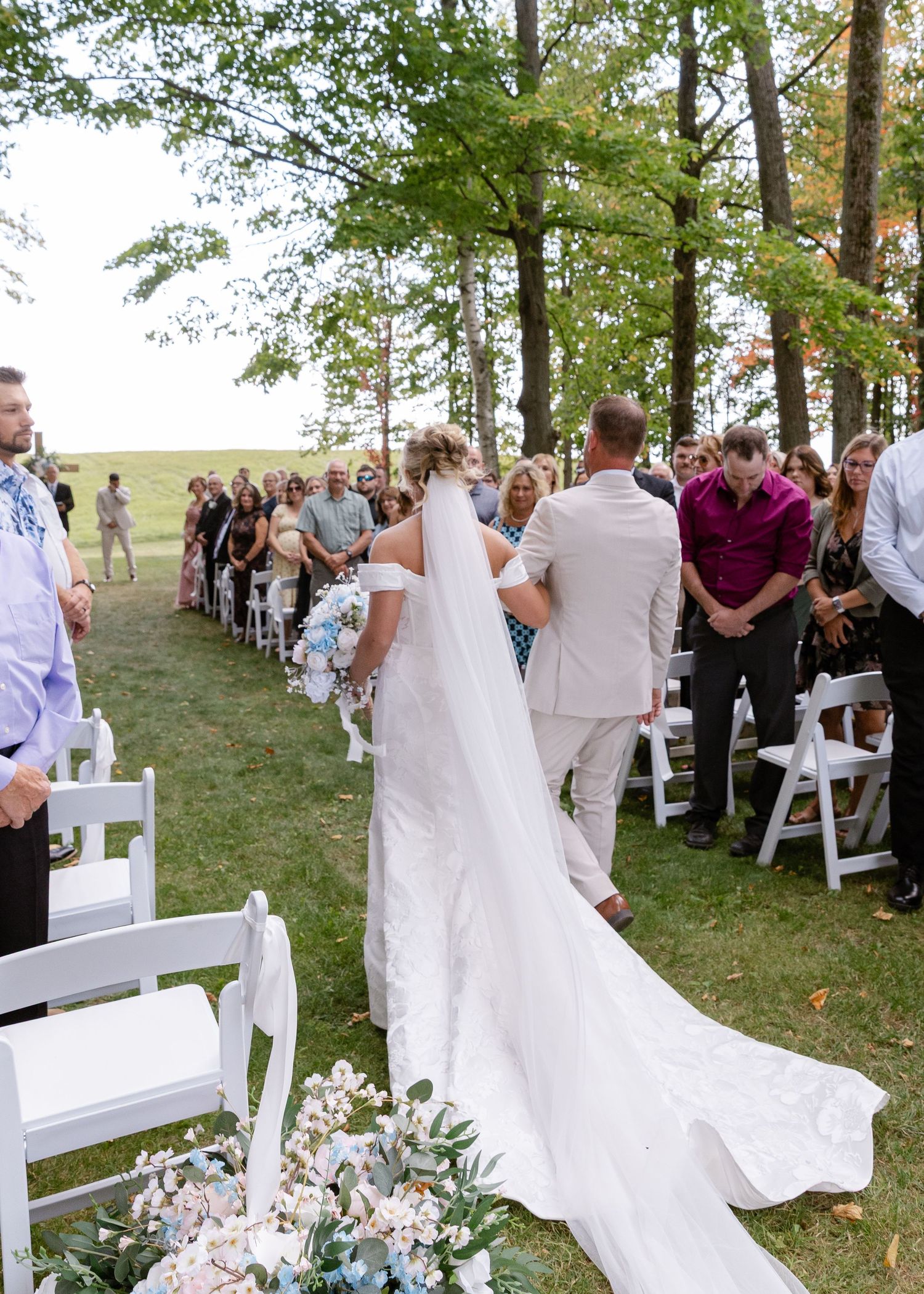 Outdoor wedding ceremony setup with white chairs and floral arrangements under tall trees.