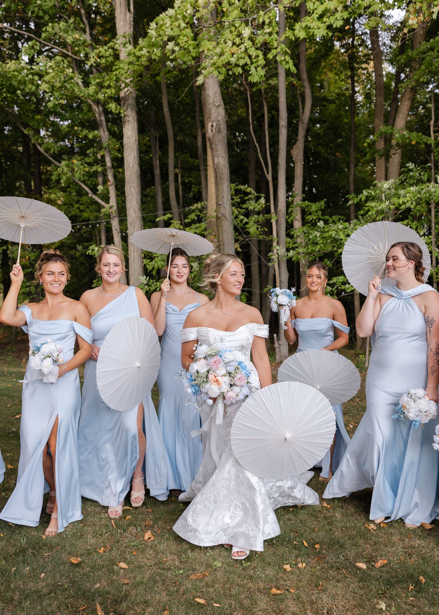 Bridesmaids in light blue dresses pose with white parasols in a wooded setting.