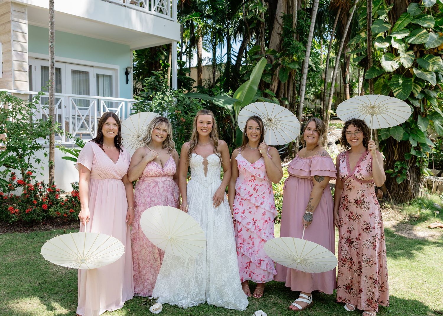 Bride in white gown poses with six bridesmaids in pink floral dresses holding parasols outdoors at a tropical venue.