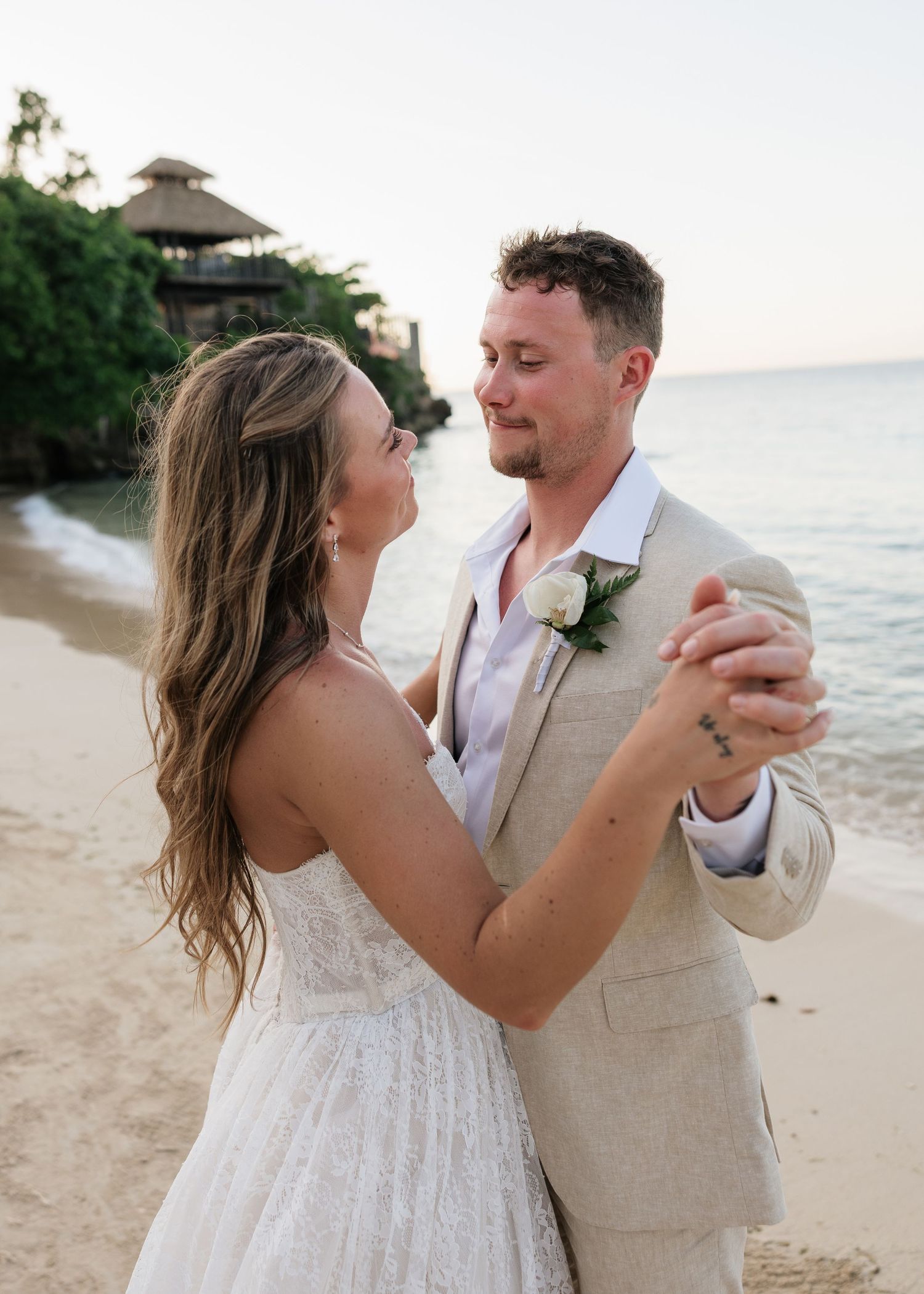 Couple sharing first dance on tropical beach at sunset, groom in tan suit with white rose boutonniere, bride in white lace gown.