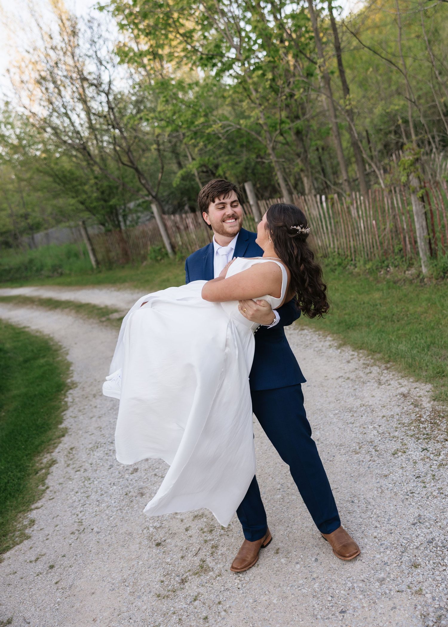 Groom in navy suit lifts smiling bride in white dress on a gravel path surrounded by green trees.
