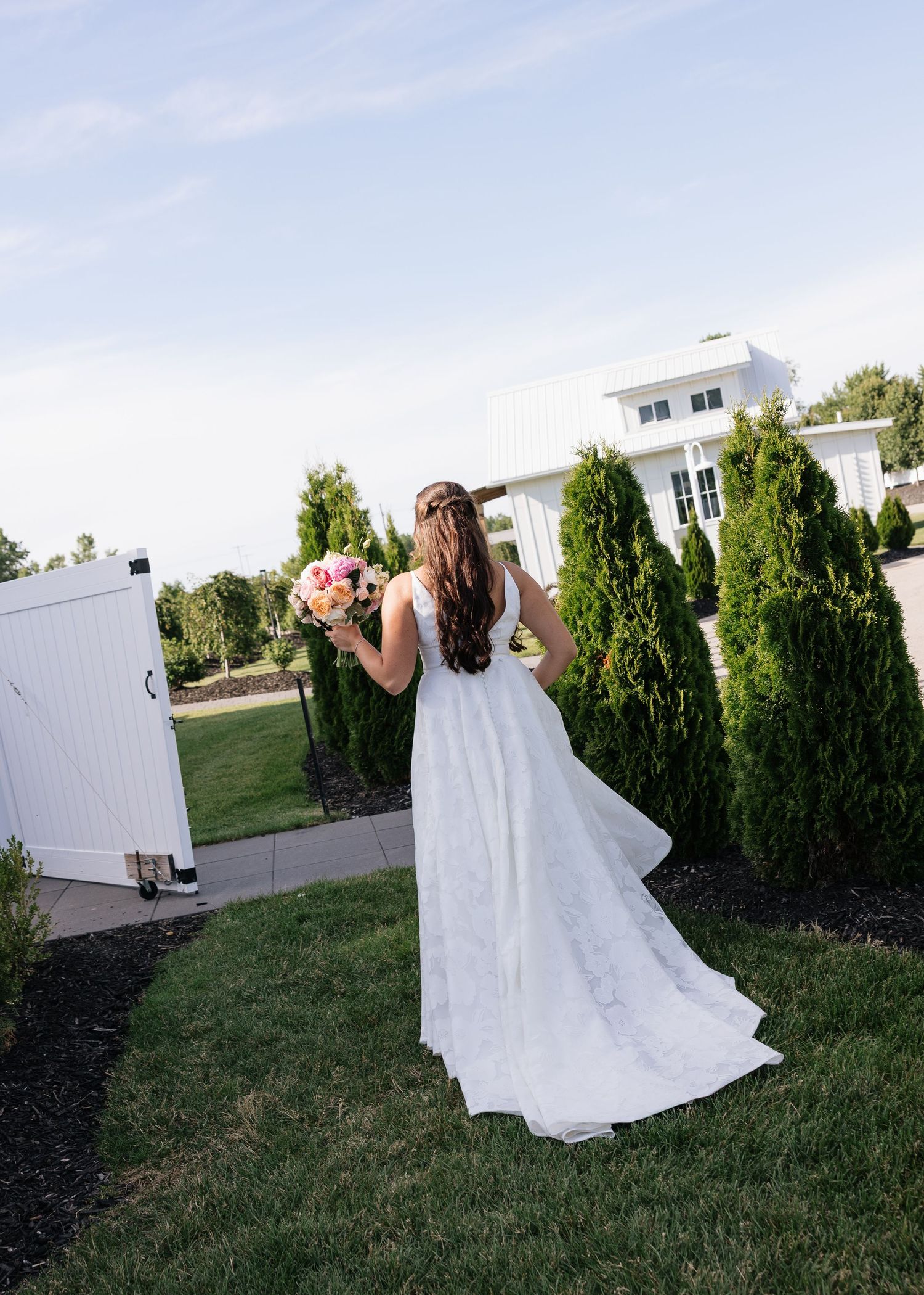 Bride in white wedding gown holding pink bouquet stands near tall hedges outside elegant white venue.