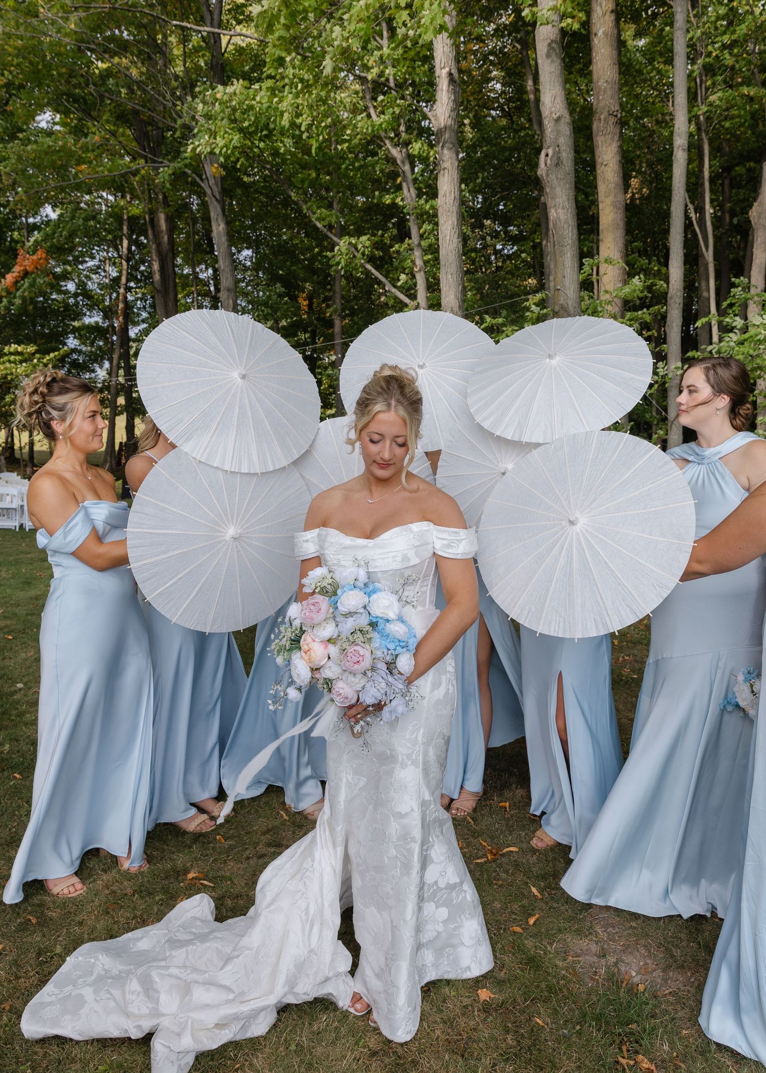 Wedding party in light blue dresses poses with white parasols against woodland backdrop.