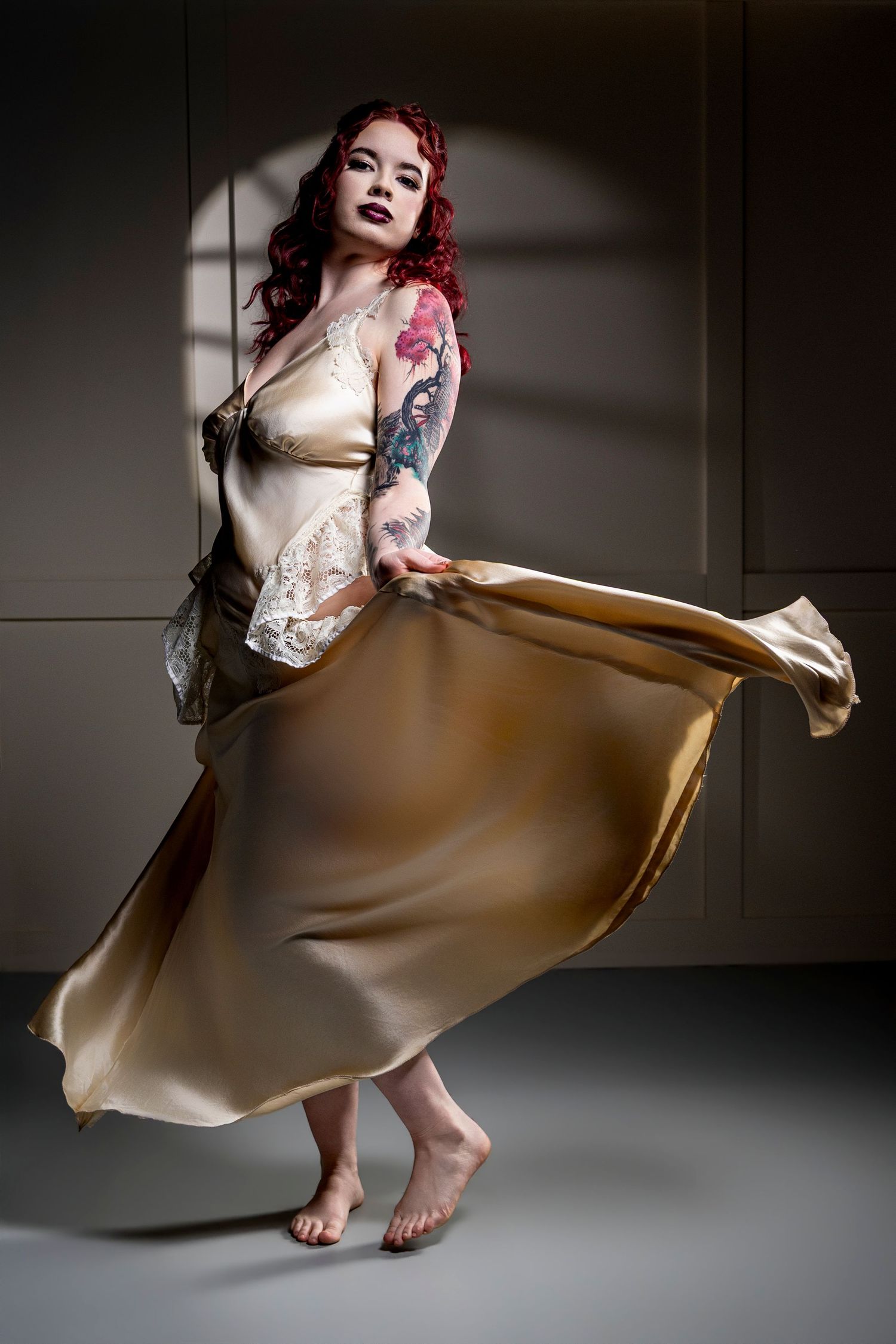 Woman with red curly hair and arm tattoos twirls barefoot in a flowing champagne satin gown with lace trim against a gray studio backdrop.