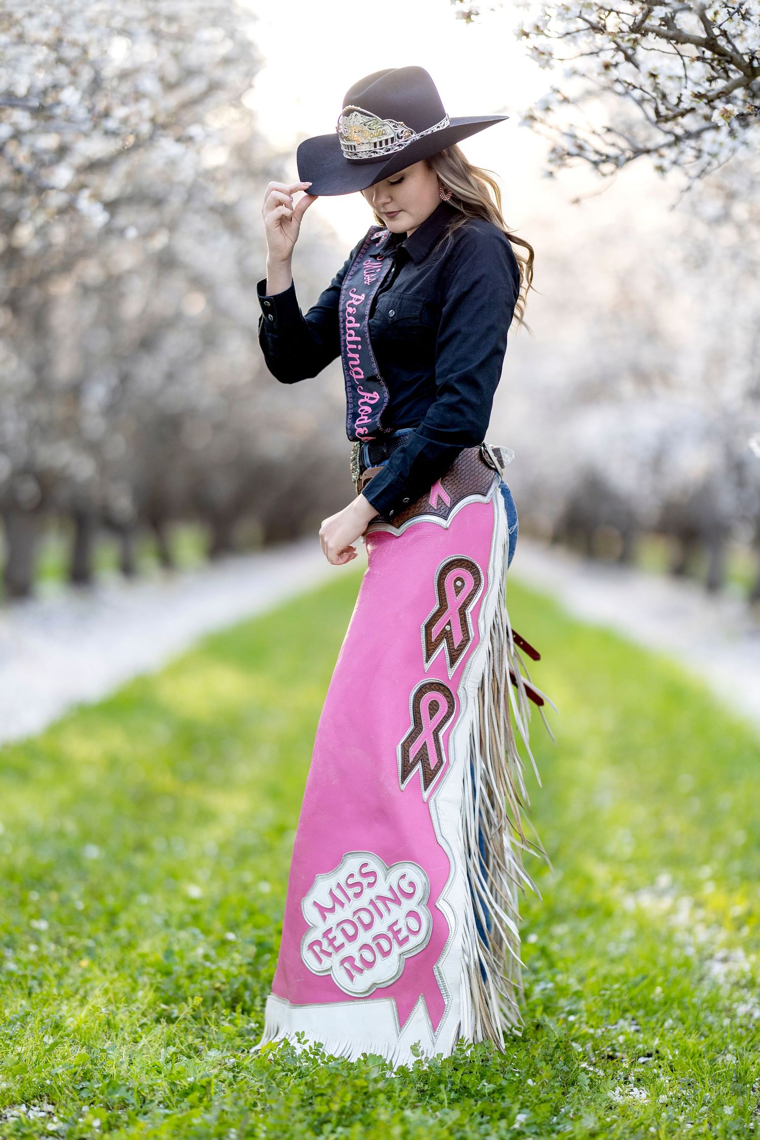Miss Redding Rodeo queen in pink fringed chaps and black cowboy hat in blooming orchard.