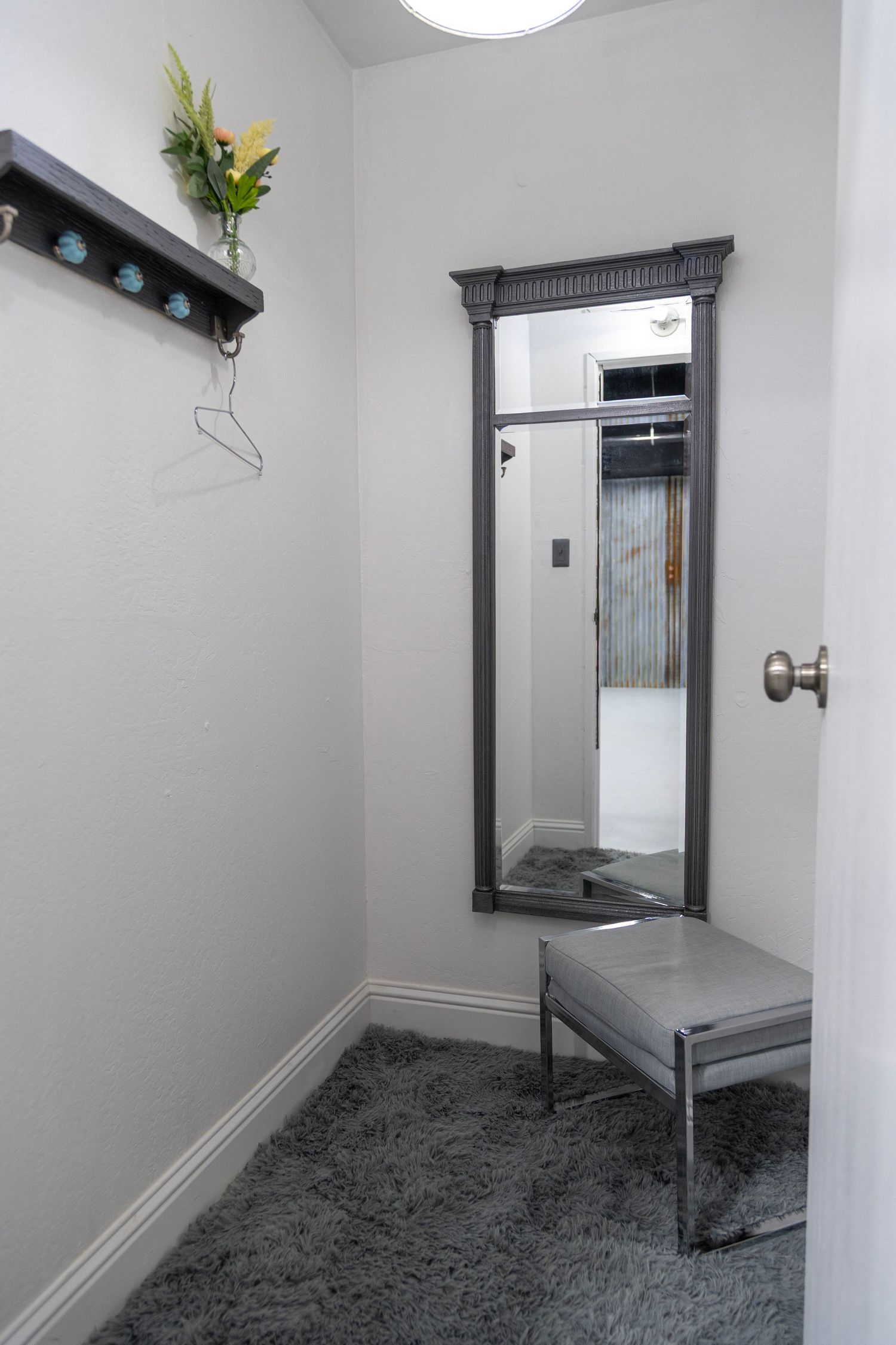 Minimalist dressing room with full-length mirror, gray bench, shag carpet, and decorative shelf with flowers.