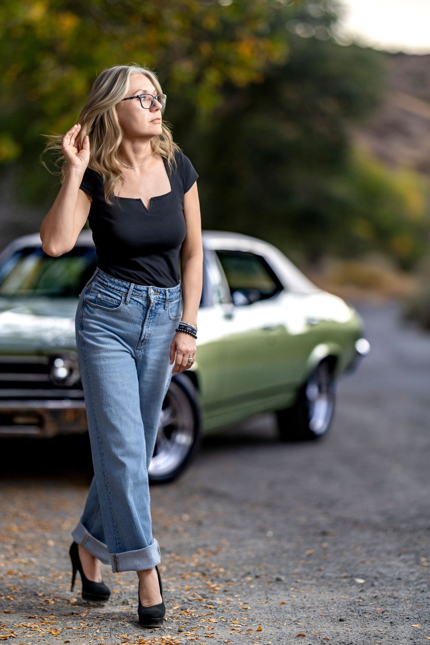 Woman with blonde hair and glasses in a black top, wide-leg jeans and heels walking past a vintage green muscle car on a rural road.