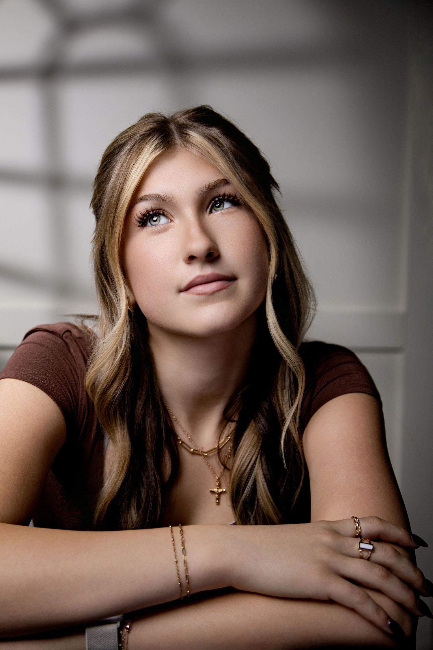 Young woman with long blonde-highlighted hair gazes upward in a portrait, wearing a brown top, gold necklaces, bracelets and rings.