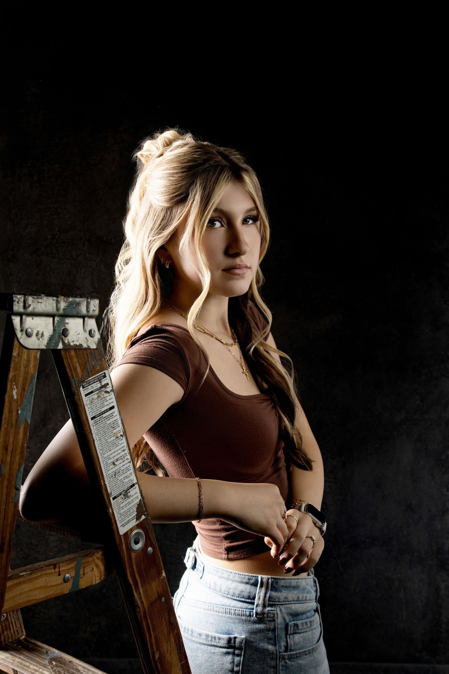 Young woman with wavy blonde hair in a half-up bun leans on a wooden ladder in a moody studio portrait against a dark backdrop.
