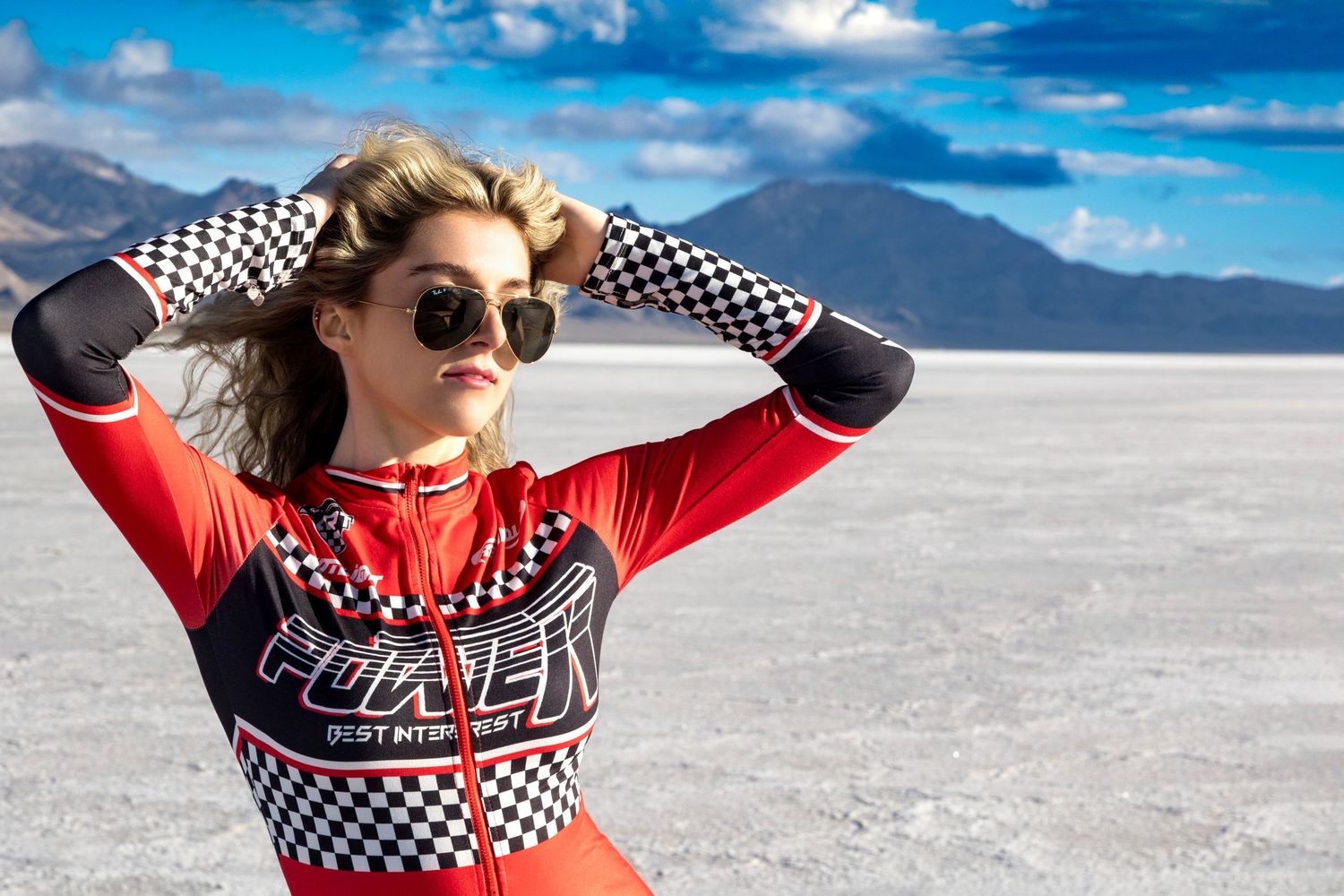 Woman in red racing suit poses on salt flats with mountains and blue sky backdrop.