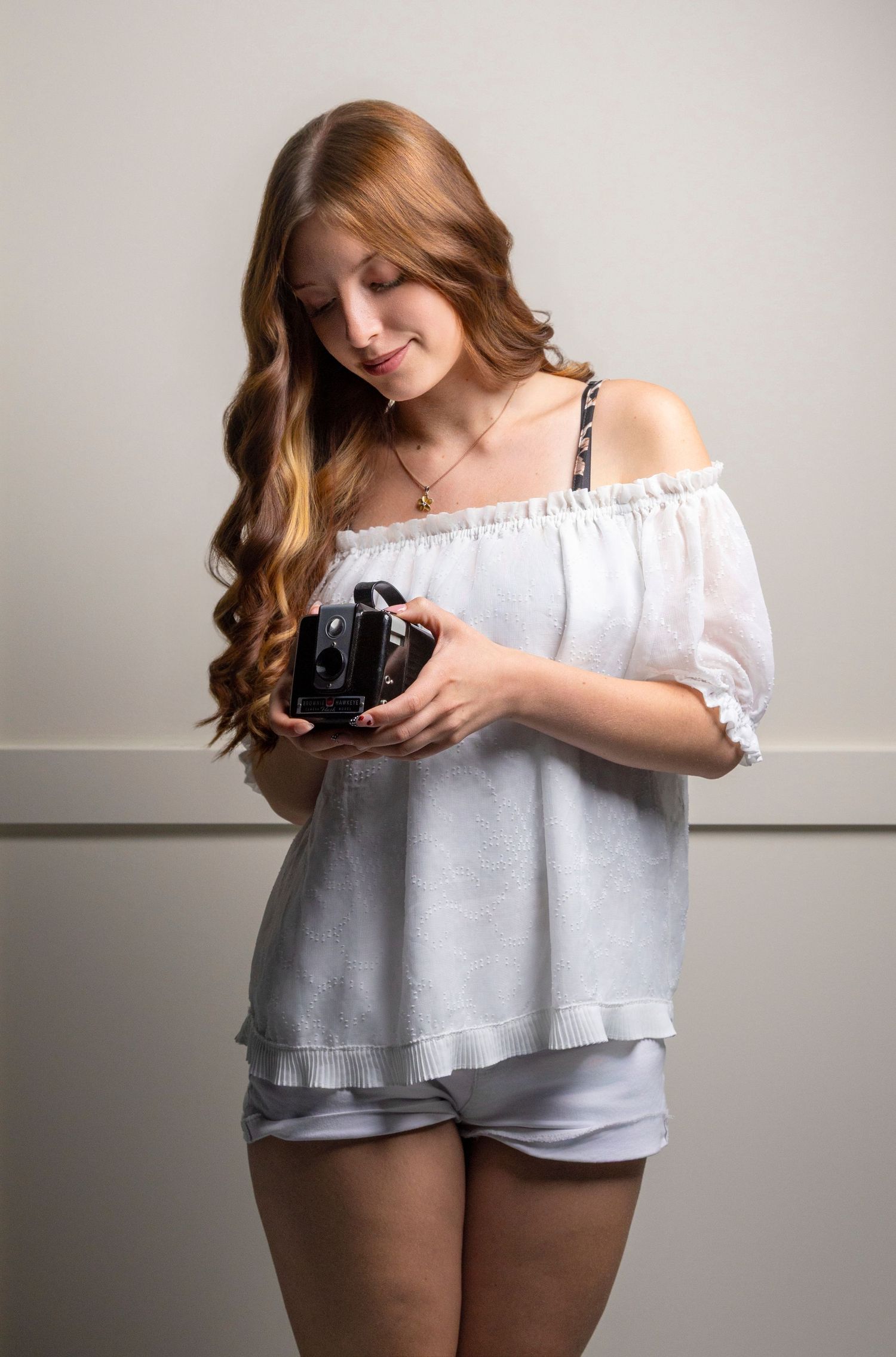 Young woman with long red wavy hair holding a vintage camera, wearing an off-shoulder white top and shorts.