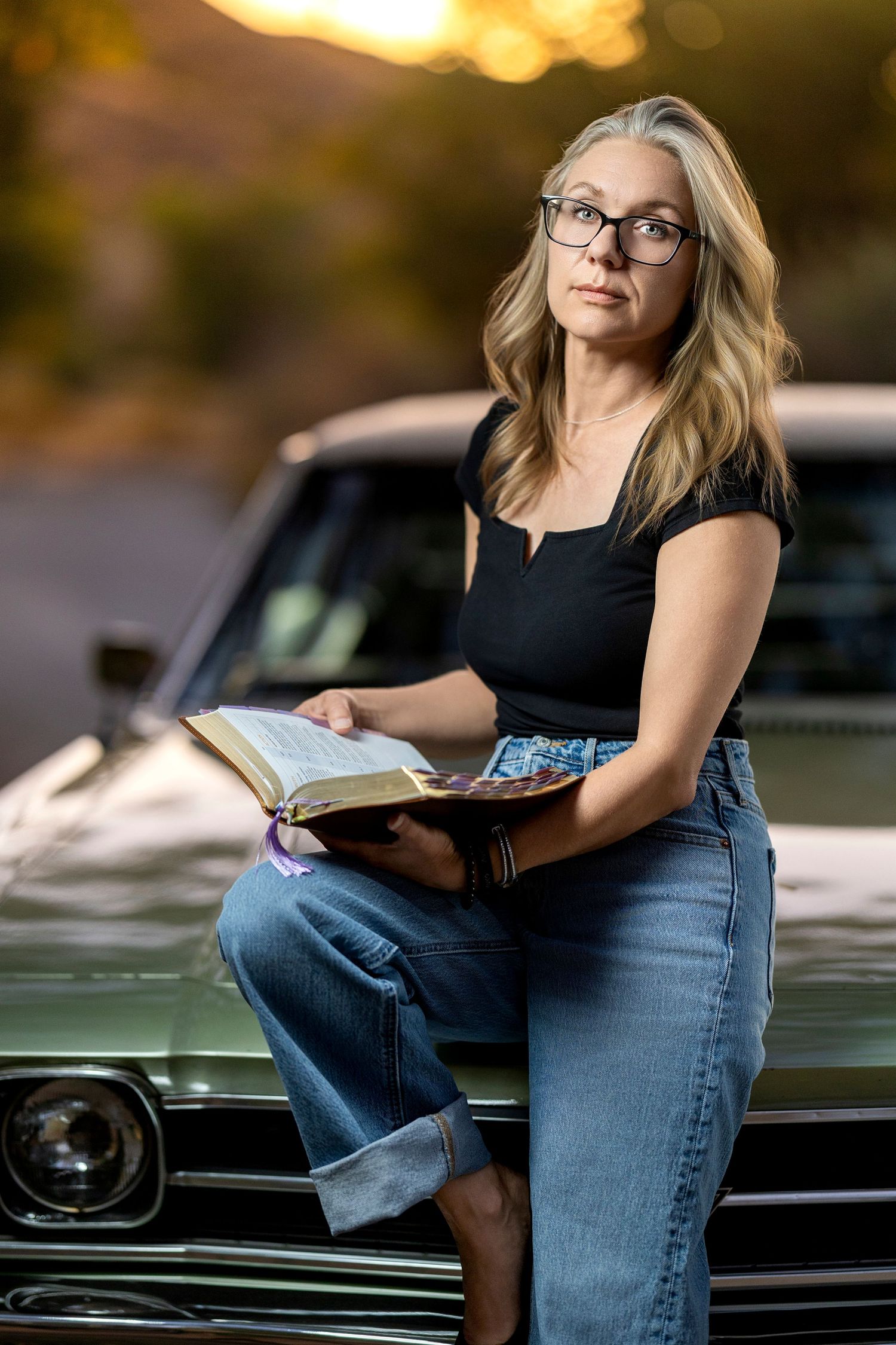 Woman with long blonde hair and glasses sits on a vintage car hood at sunset, holding an open Bible in cuffed jeans.