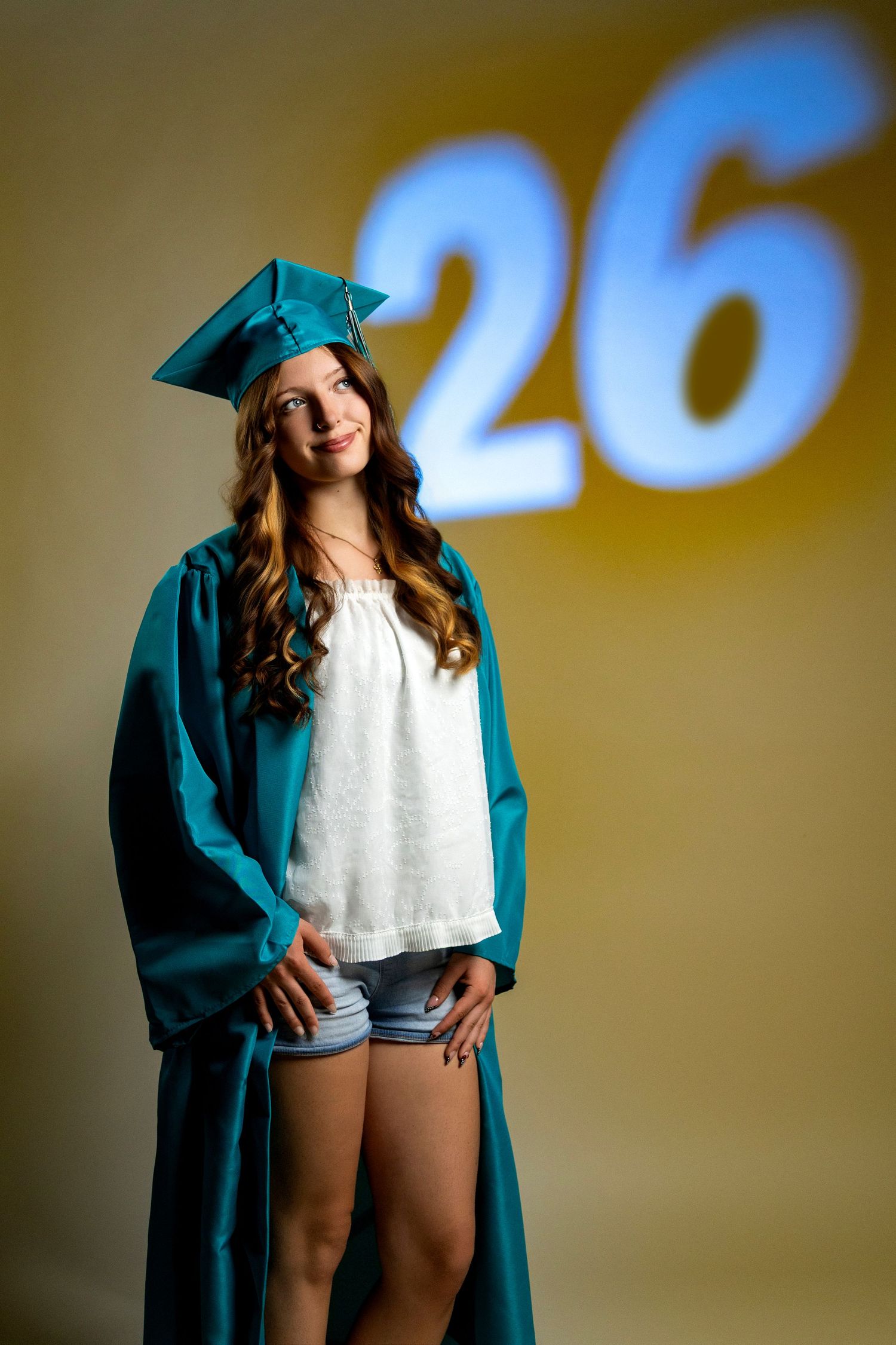 Smiling teen girl in teal graduation cap and gown posing in front of a projected number 26 background.