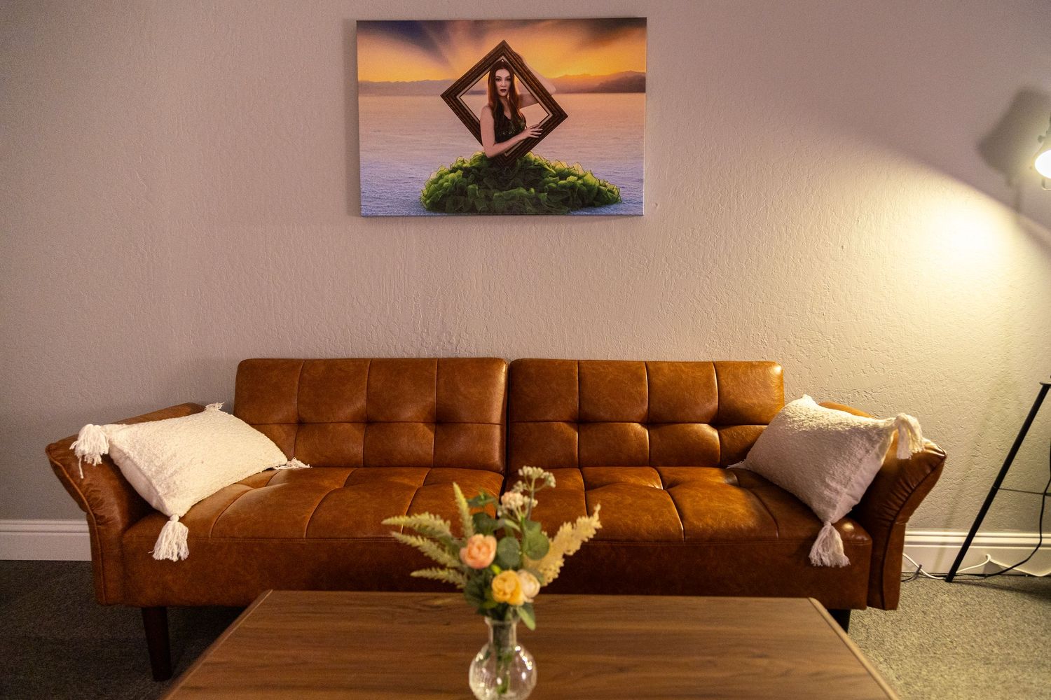 Brown leather tufted sofa with white pillows below a canvas art print, with a wood coffee table and flower vase.