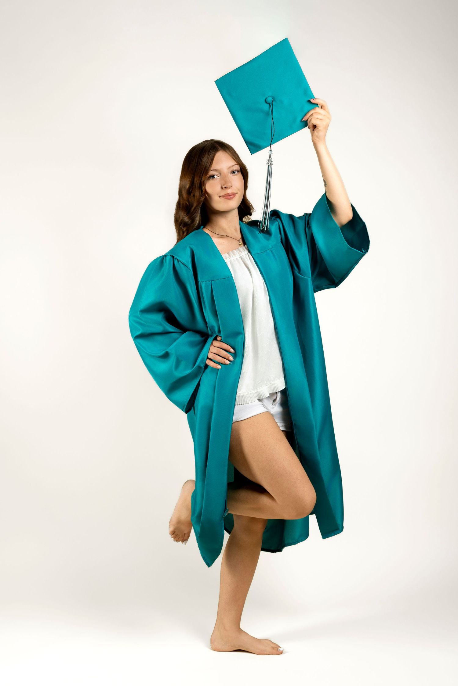 Teen girl in teal graduation gown and cap raised overhead, posing barefoot on a white studio background.