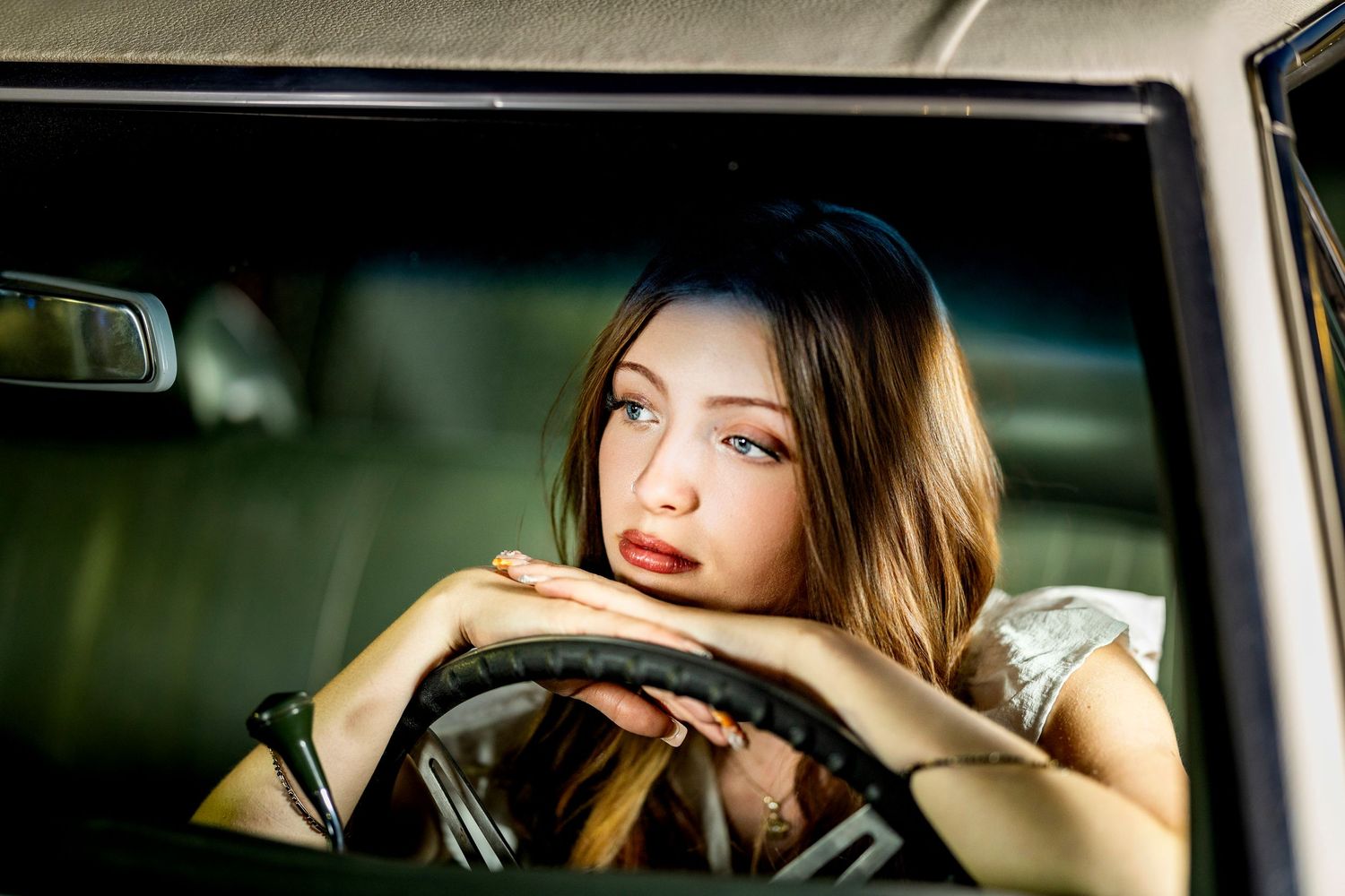 Young woman with long hair resting her chin on hands over a steering wheel inside a vintage car interior.