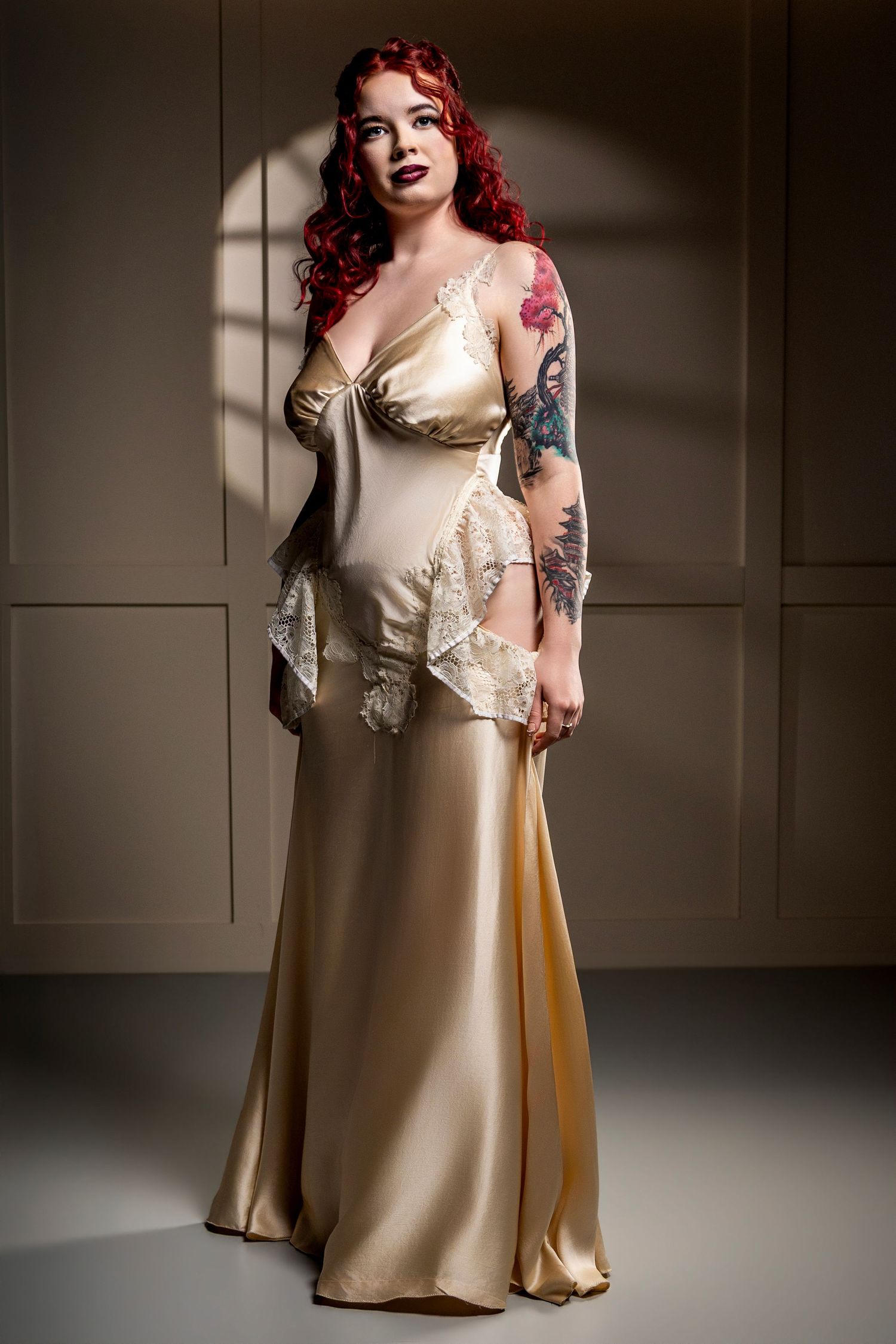 Woman with curly red hair and arm tattoos poses in a champagne satin gown with lace trim against a neutral paneled studio wall.