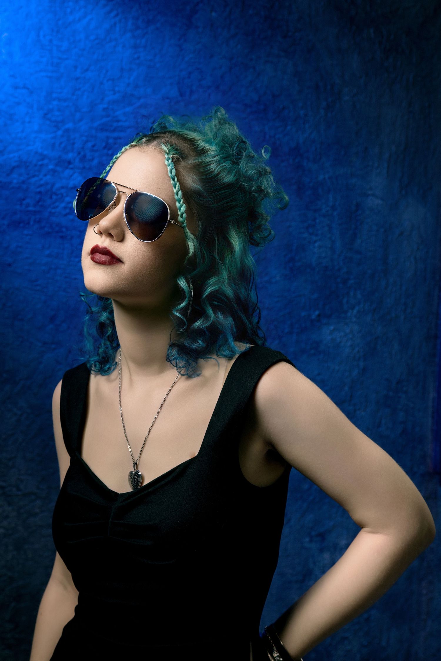 Young woman with teal curly hair wearing sunglasses and black dress poses confidently against blue background.