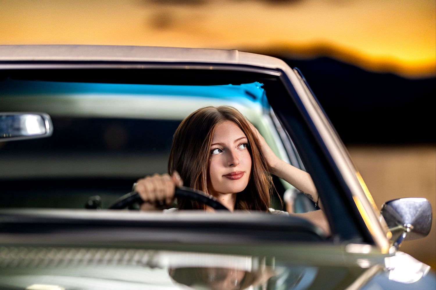 Young brunette woman smiling in a classic car at sunset, with golden hour light reflecting in the background.