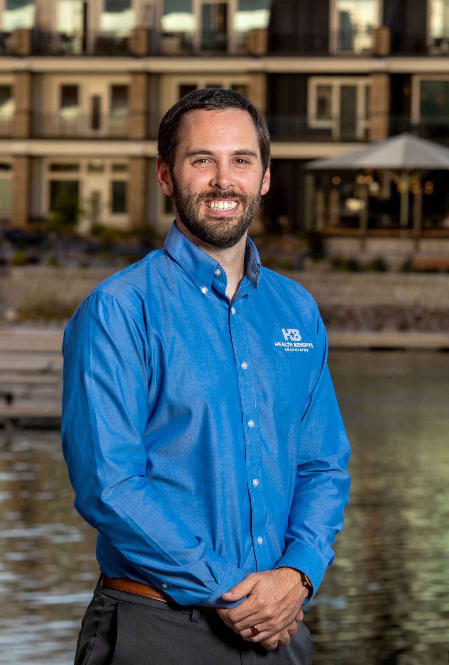 Smiling man in blue button-up shirt standing outdoors near waterfront building on a sunny day.