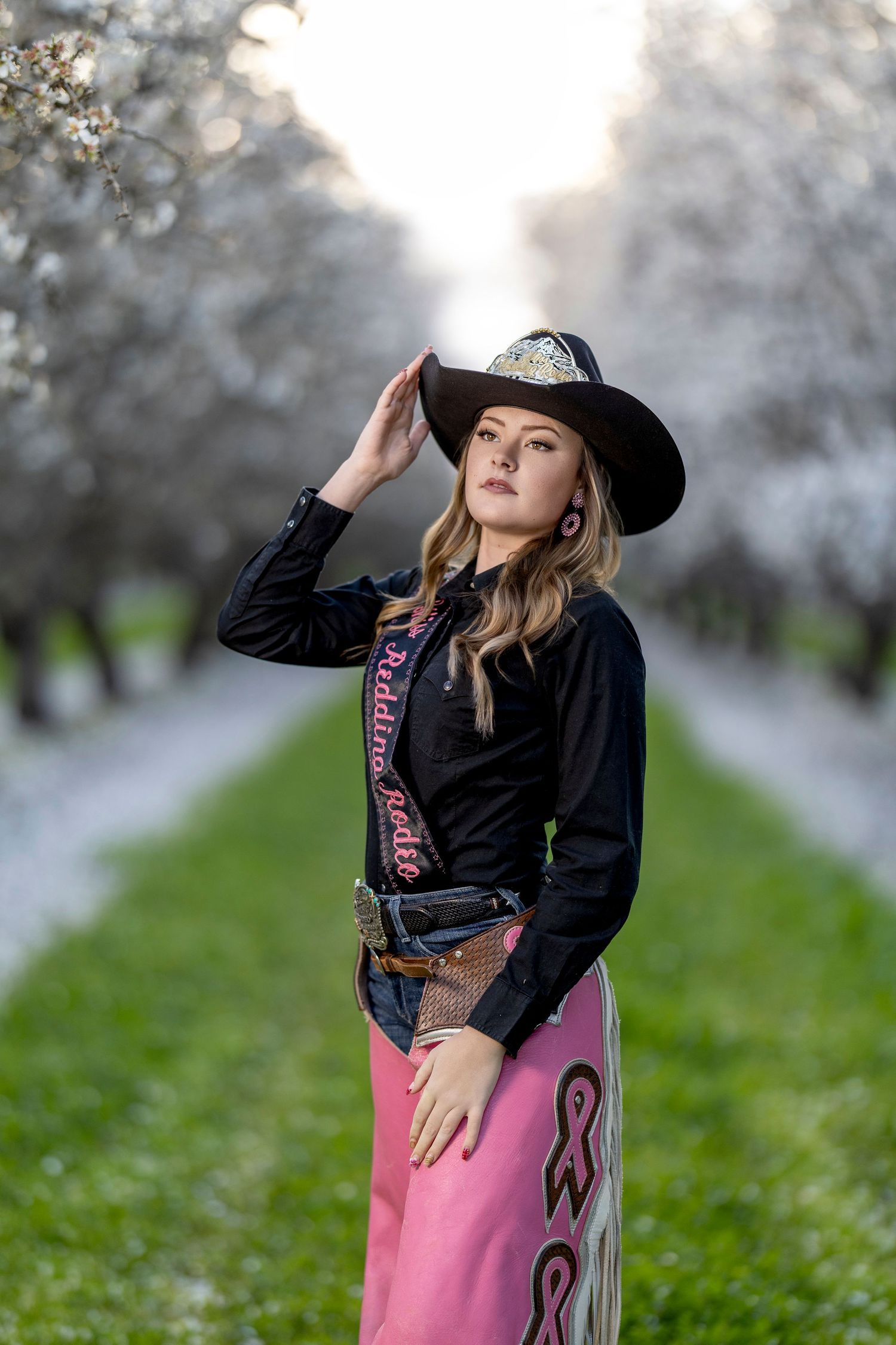 Miss Redding Rodeo contestant in black shirt, pink chaps, and cowboy hat in orchard.