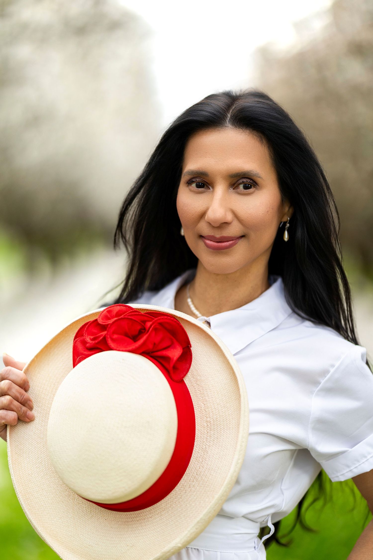 Dark-haired woman holding straw hat with red roses in sunlit blossoming garden path.