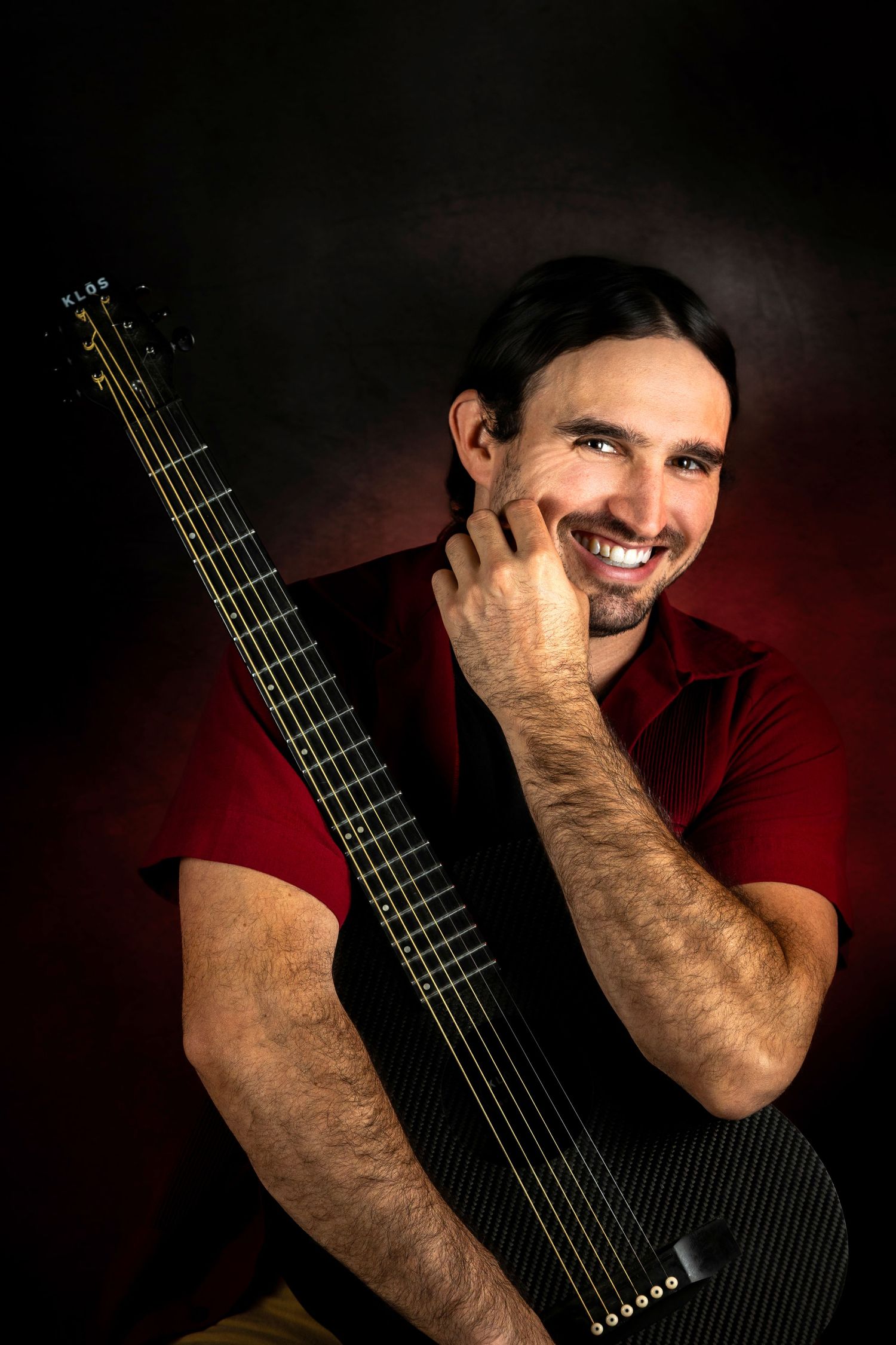 Smiling man with dark hair holding a black multi-string guitar against a red dark background.