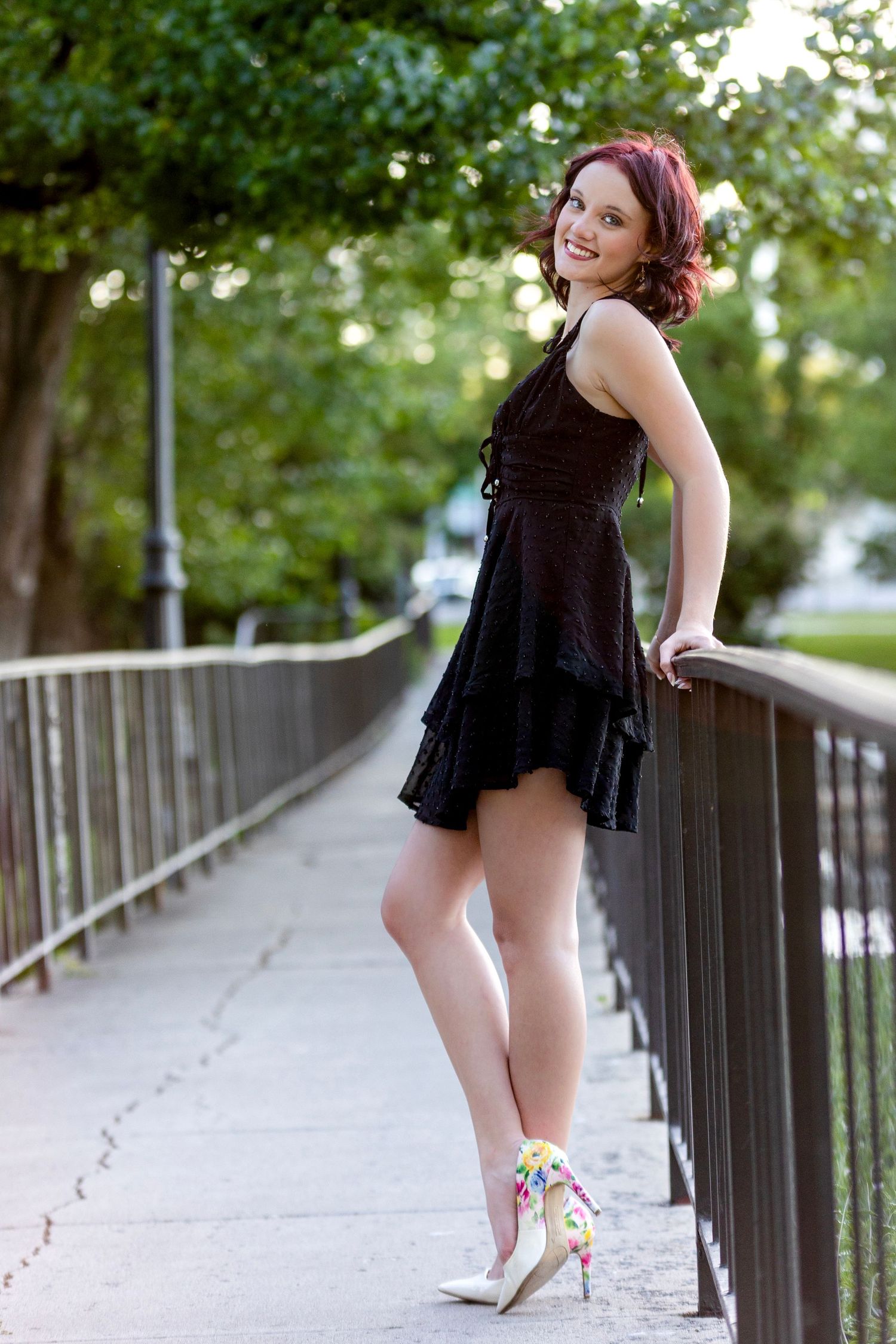 Smiling redhead woman in a black mini dress leans on a railing on a bridge outdoors.