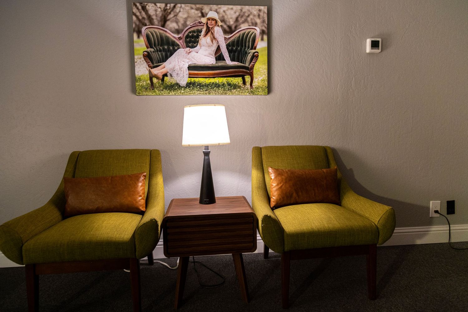 Two green mid-century modern chairs with orange pillows flank a table lamp beside a portrait photo on a gray wall.