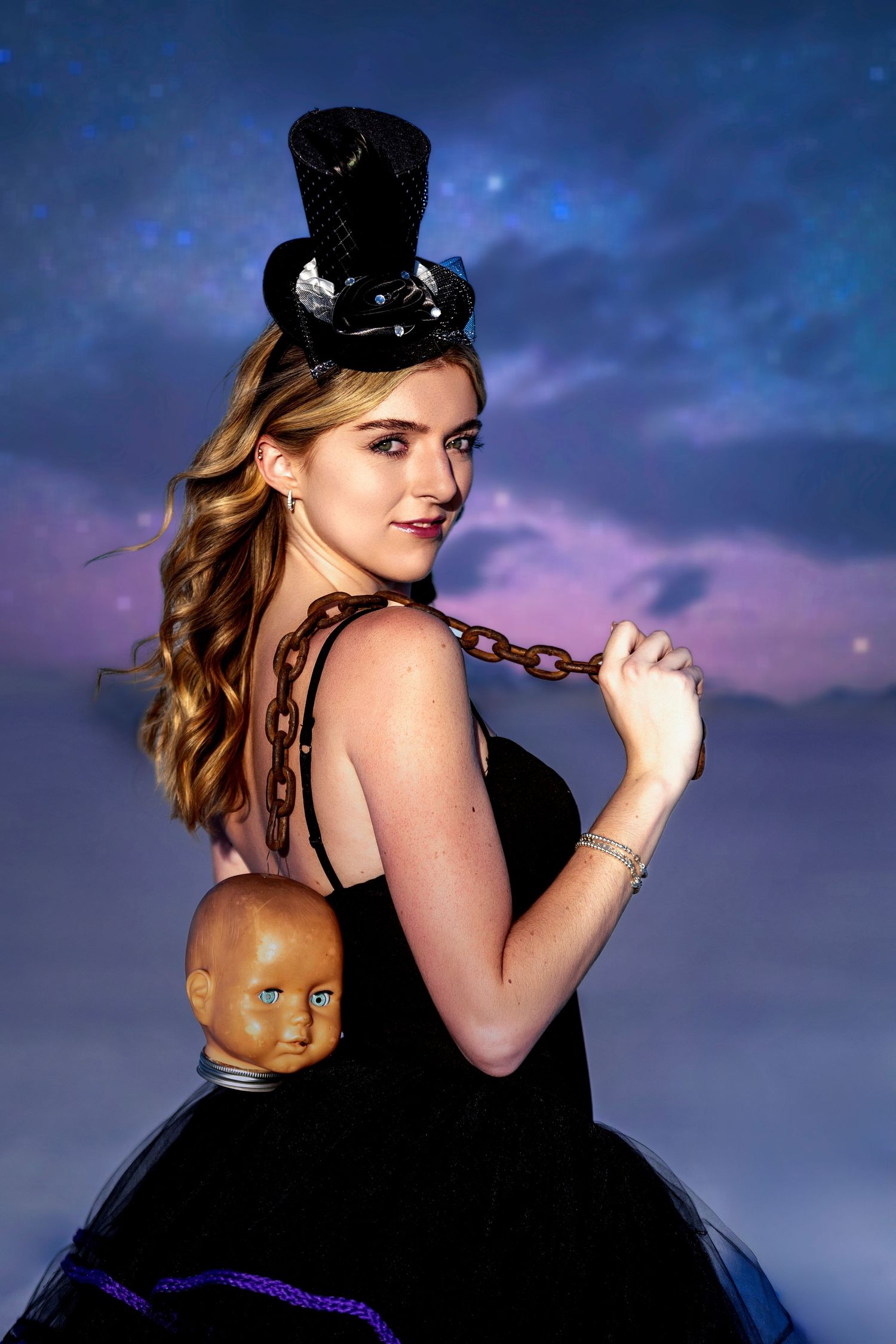 Young woman in black top hat and tutu holds doll head with chain against night sky.