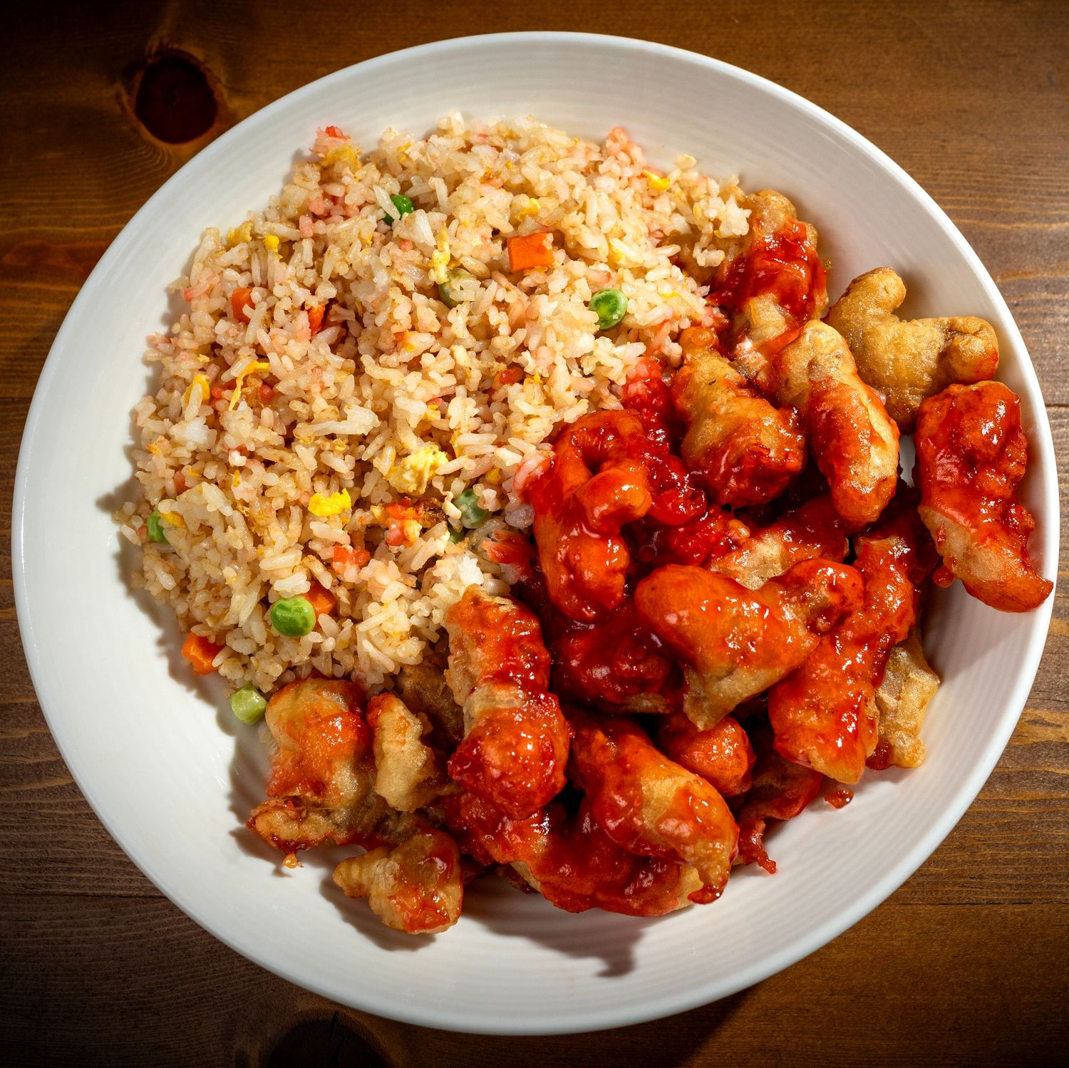 White plate of sweet and sour chicken with fried rice on wooden table.