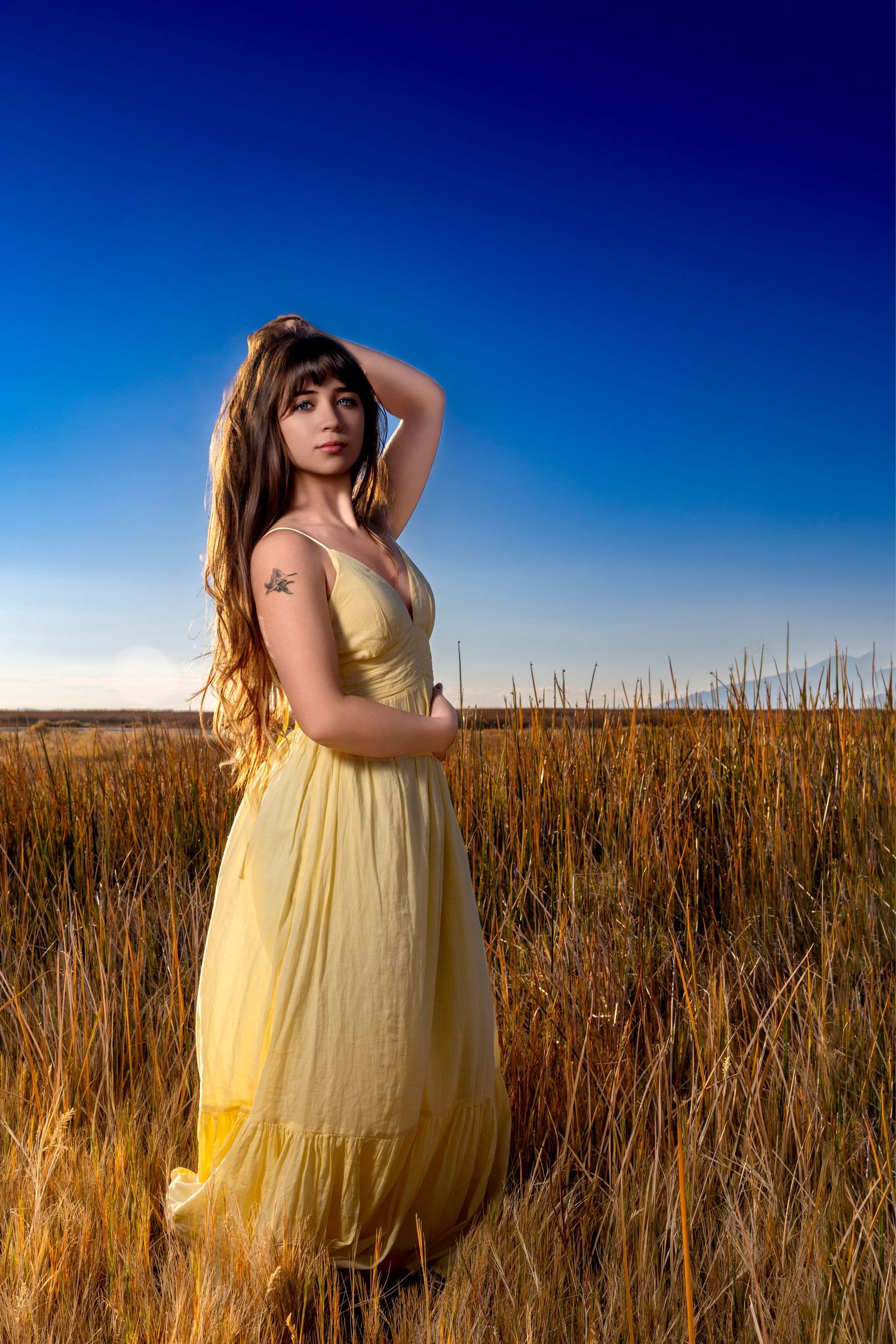 Young woman in a flowing yellow dress stands in a golden field under a deep blue sky.