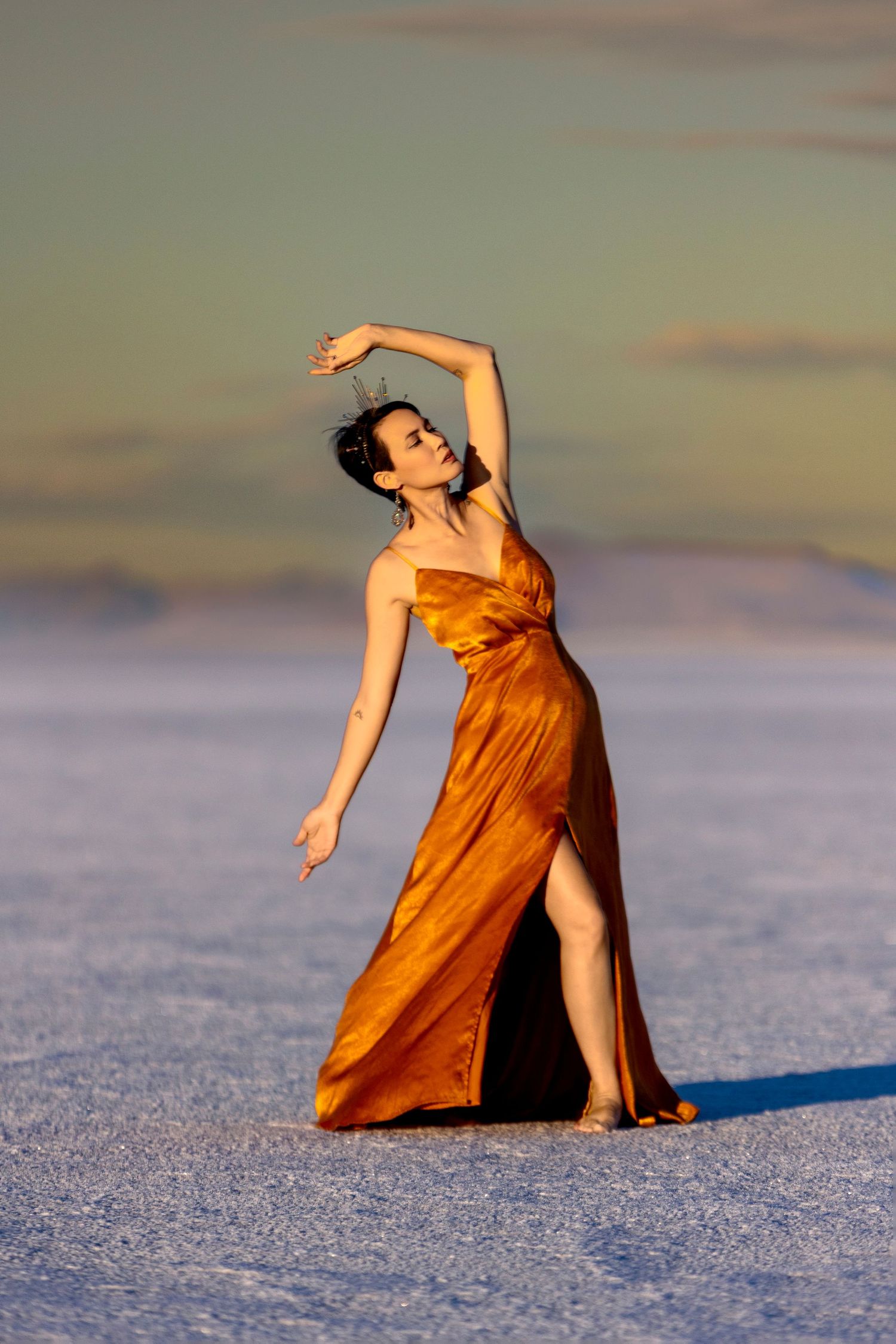 Elegant woman in gold satin gown poses gracefully on salt flats at sunset.