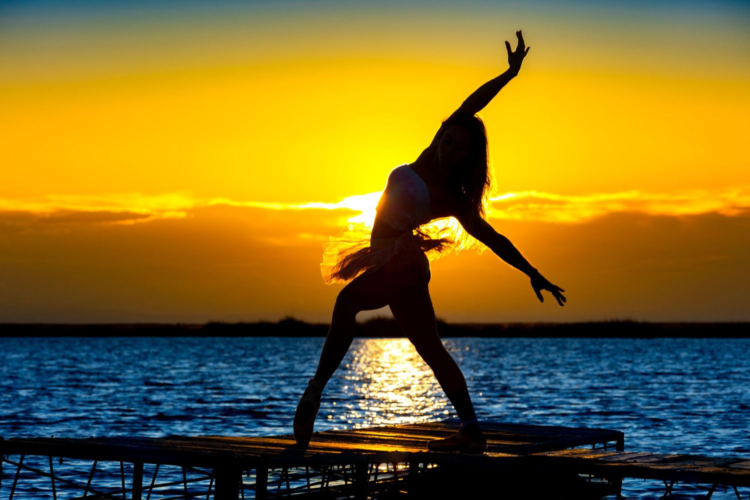 Silhouette of a dancer posing gracefully on a pier during a vibrant golden sunrise over water.
