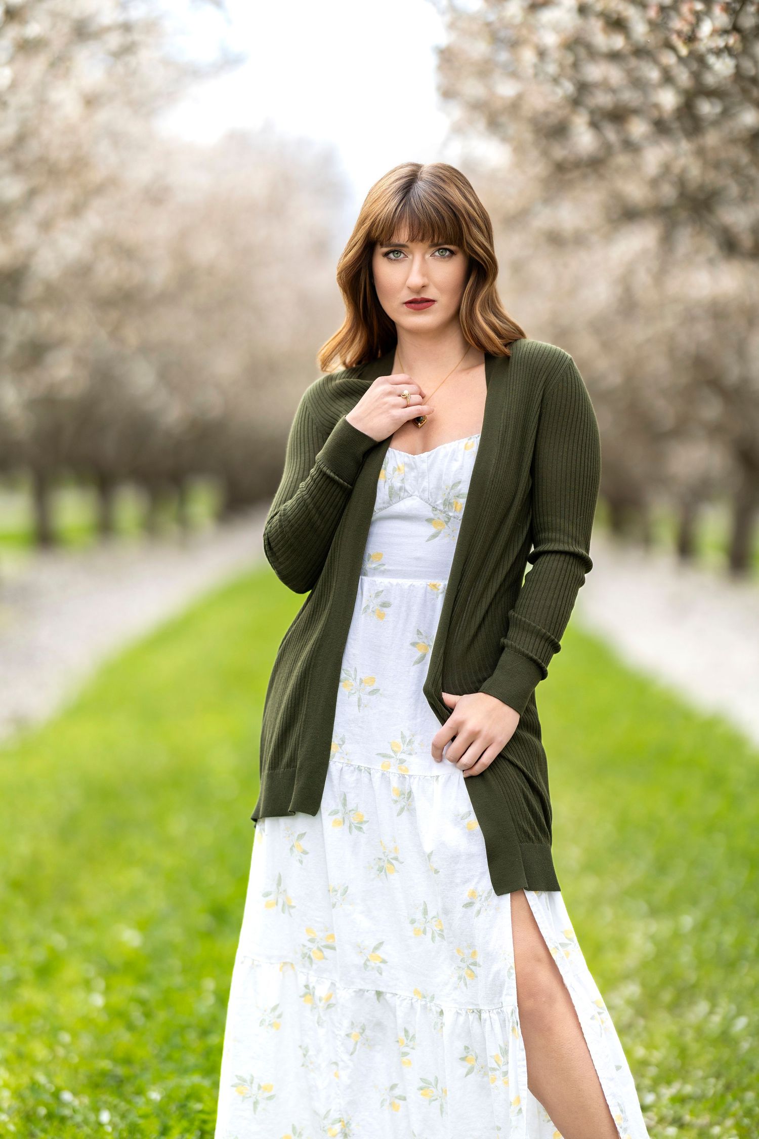 Woman in floral dress and olive cardigan posing in blooming spring orchard pathway.