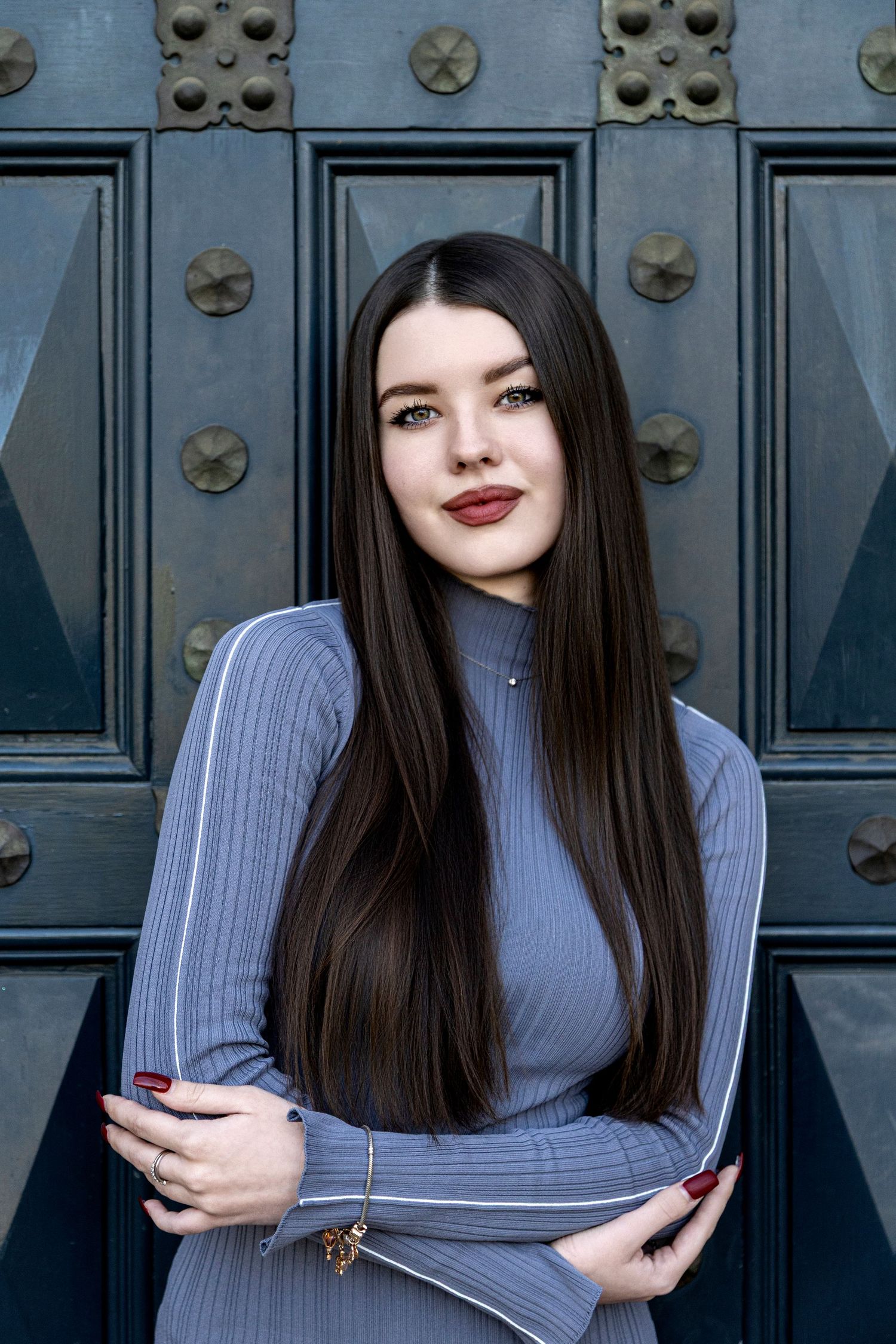Young brunette woman in gray ribbed dress poses confidently against ornate dark metal door with decorative studs.