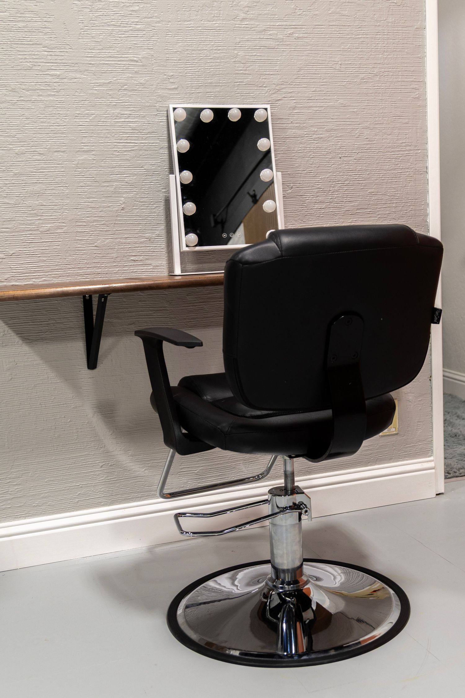 Professional black hydraulic styling chair at a vanity desk with Hollywood bulb mirror in a salon studio.