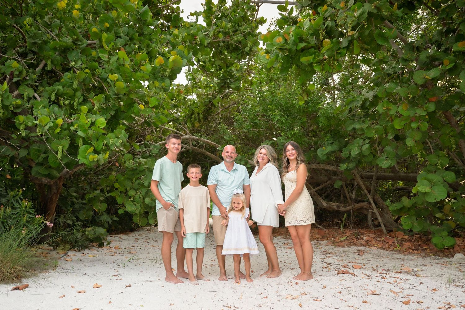 Family of six posing together on white sand beach surrounded by lush green tropical trees.