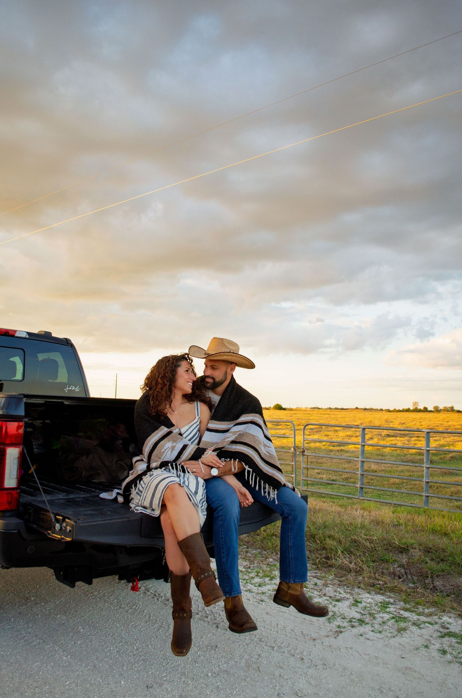 A couple sits together on the tailgate of a pickup truck at sunset in a rural setting with fields and fences in the background.