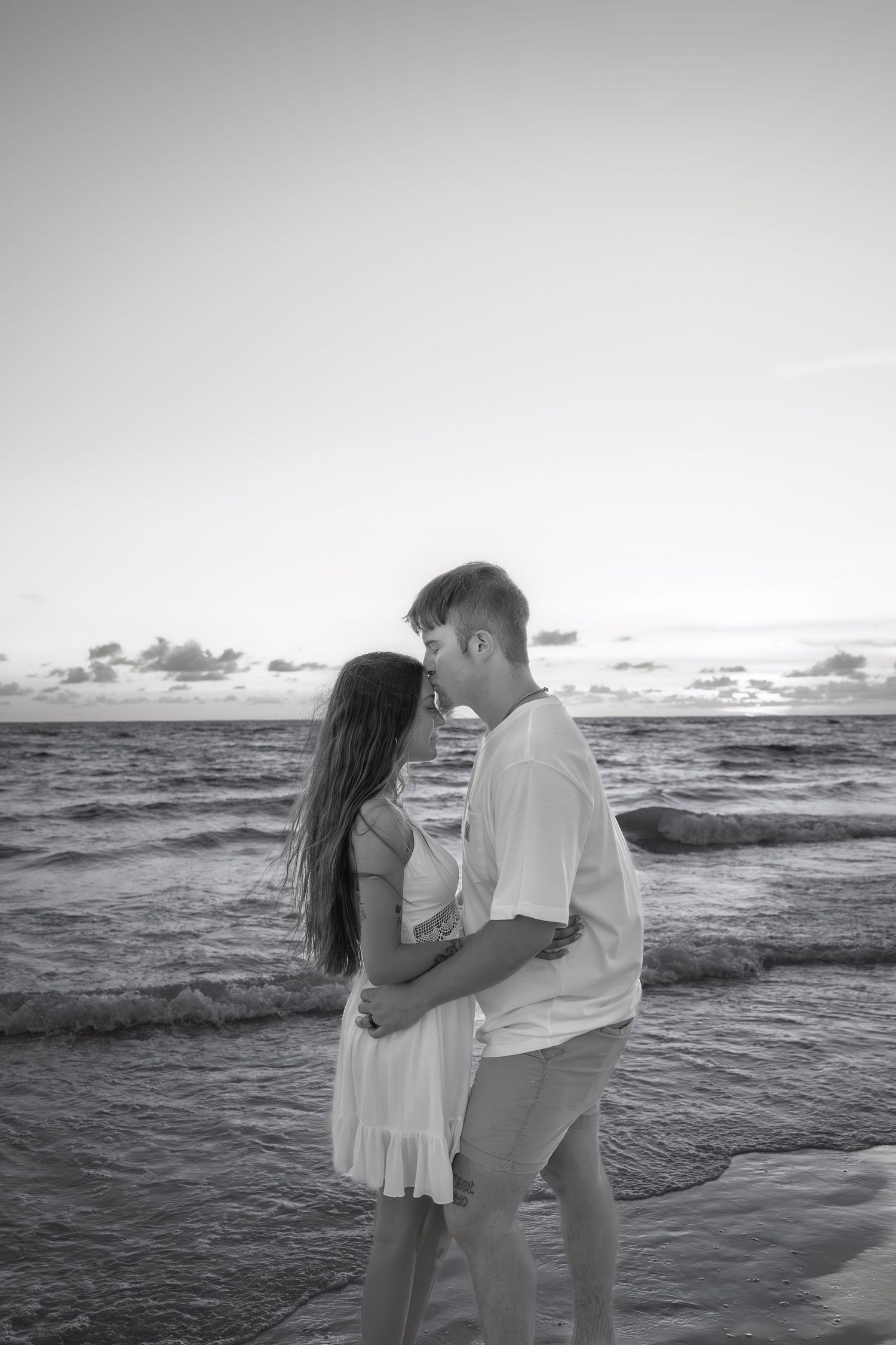 Black and white photo of romantic couple hugging on beach at sunset with ocean waves behind them.