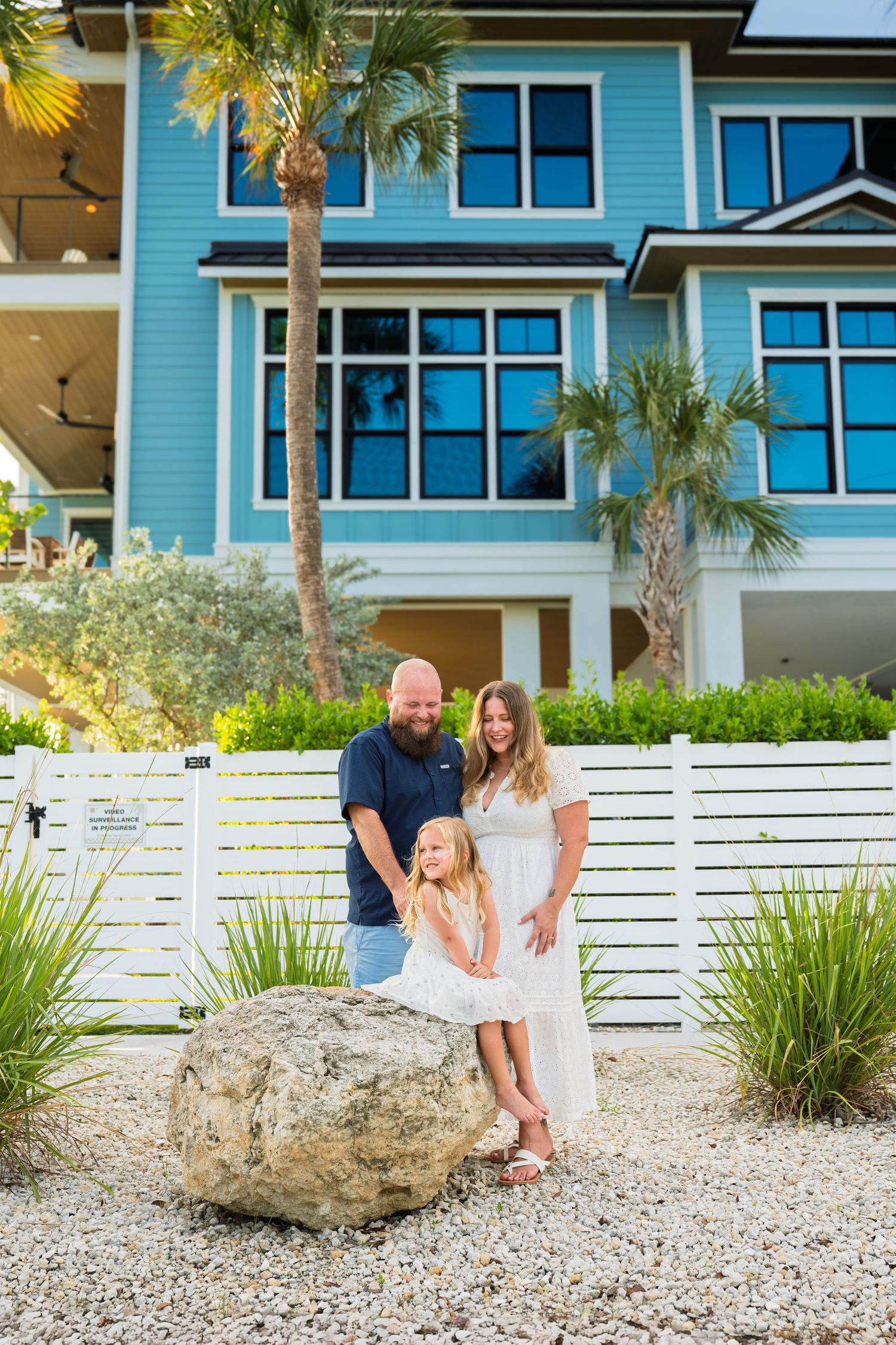 Family of three posing in front of a blue beach house with palm trees and white fence on a sunny day.