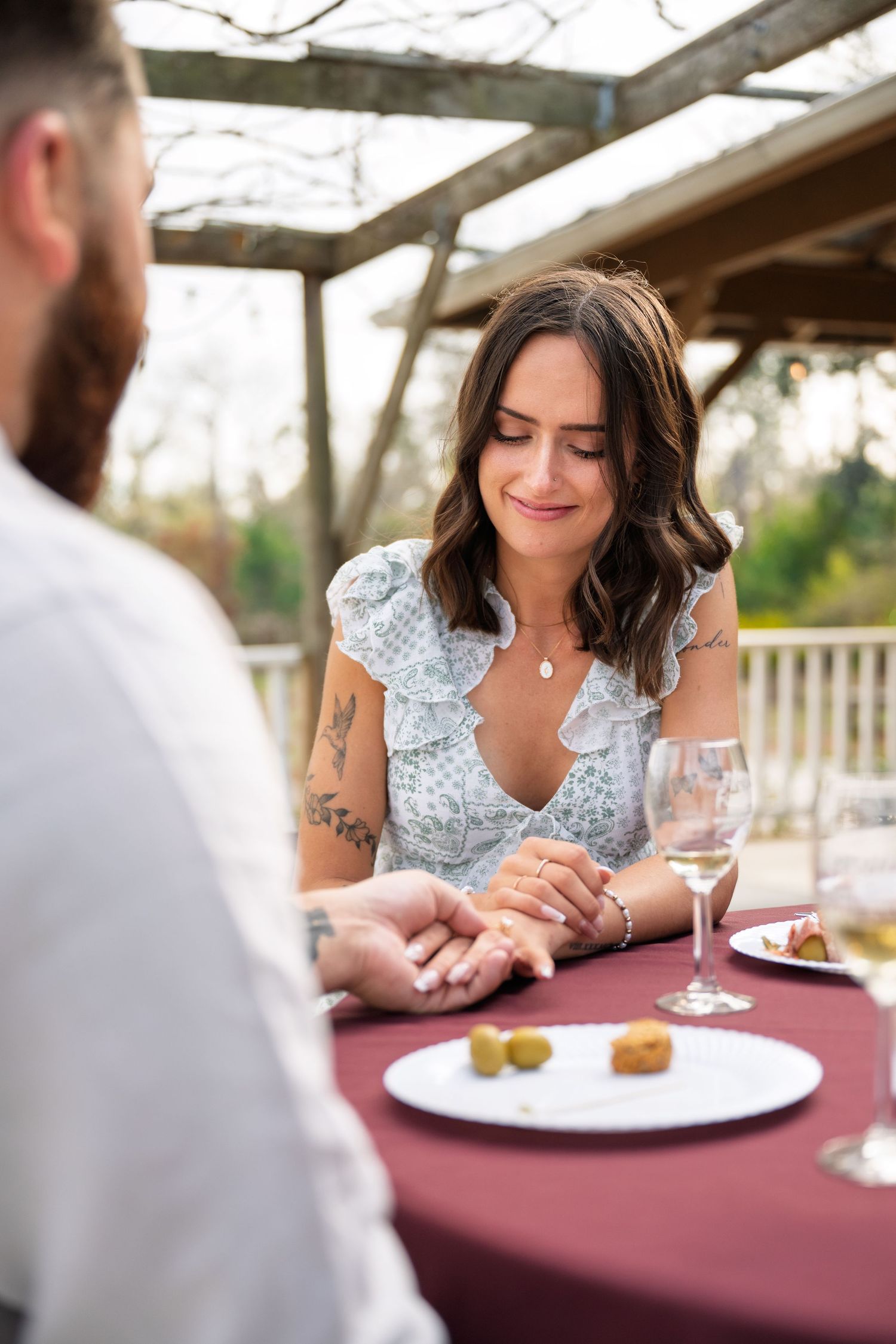 Emotional woman smiles at engagement ring while dining outdoors with partner at romantic restaurant table.