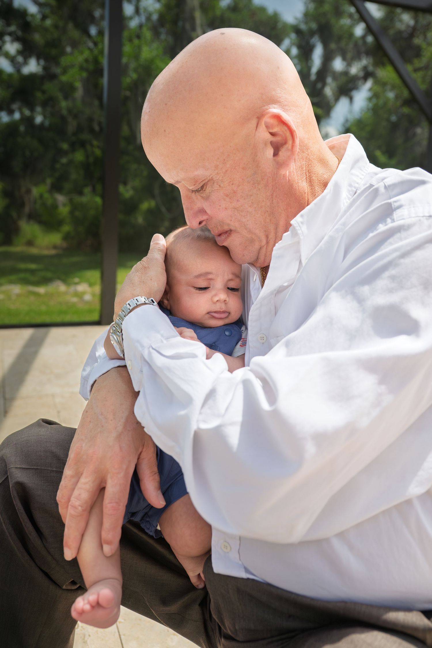 Bald grandfather tenderly holding a sleeping baby outdoors, wearing a white shirt and watch.