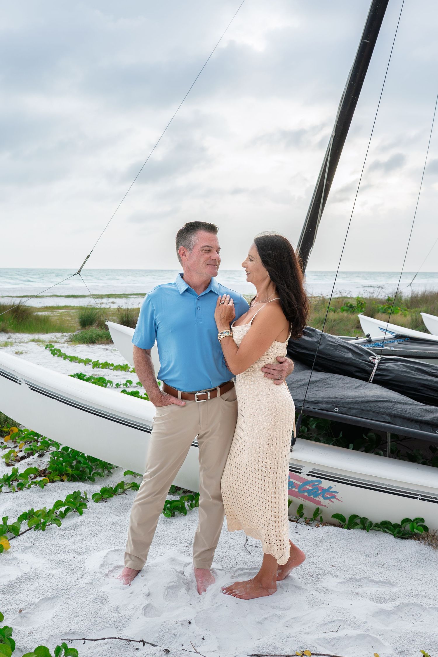 Couple embracing on white sand beach beside a sailboat, man in blue polo and woman in cream crochet dress.