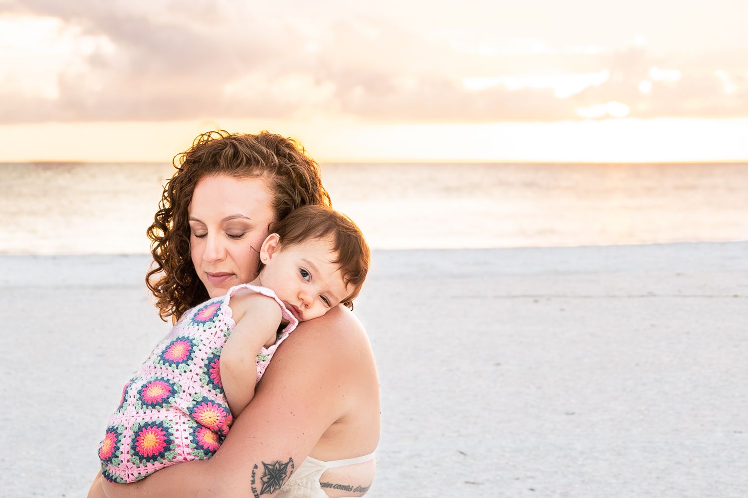 A mother tenderly holds her baby on a beach at sunset, both dressed in summer attire with ocean waves behind them.